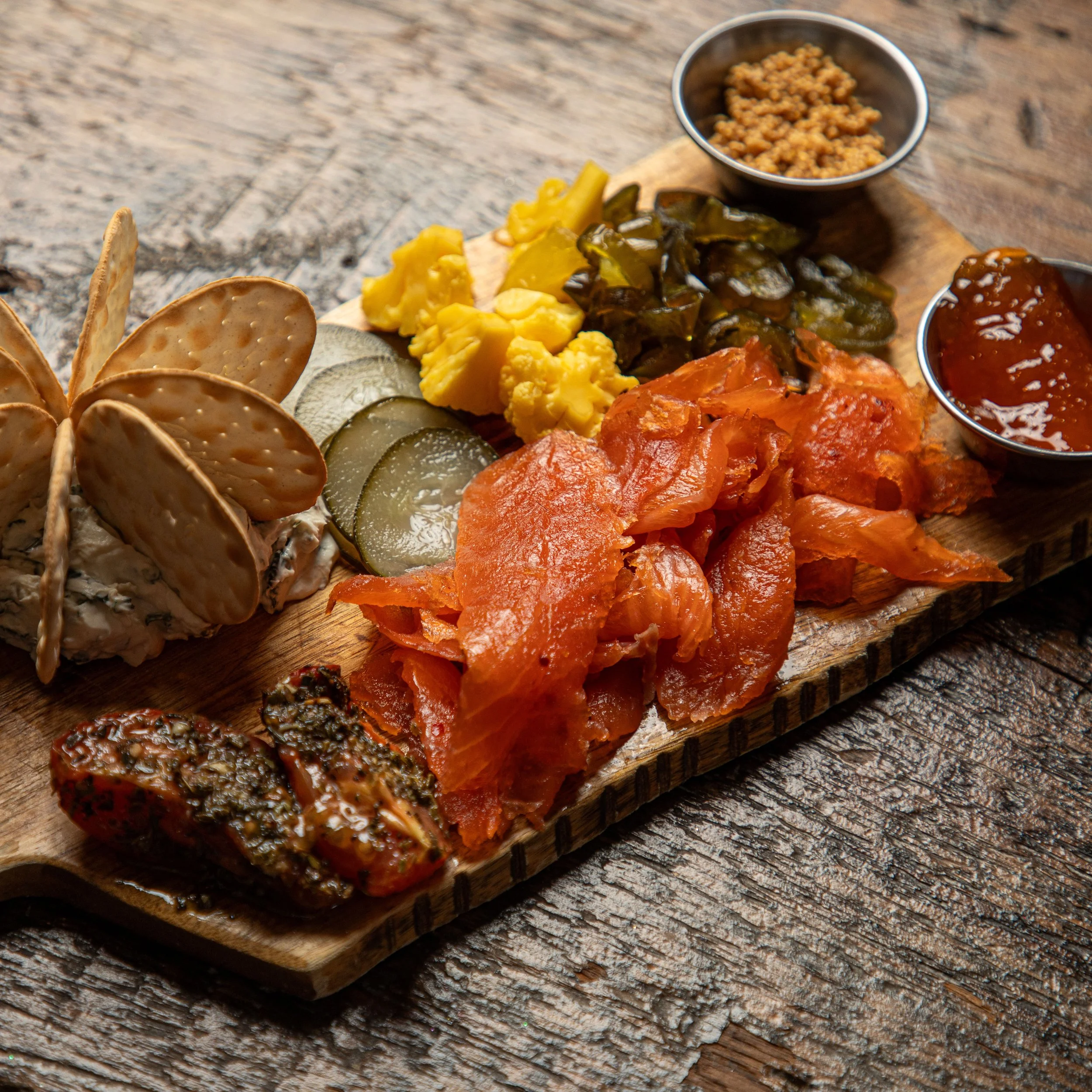 Charcuterie board with smoked salmon, pickles, yellow mustard, olives, chutney, and crackers on a wooden surface.