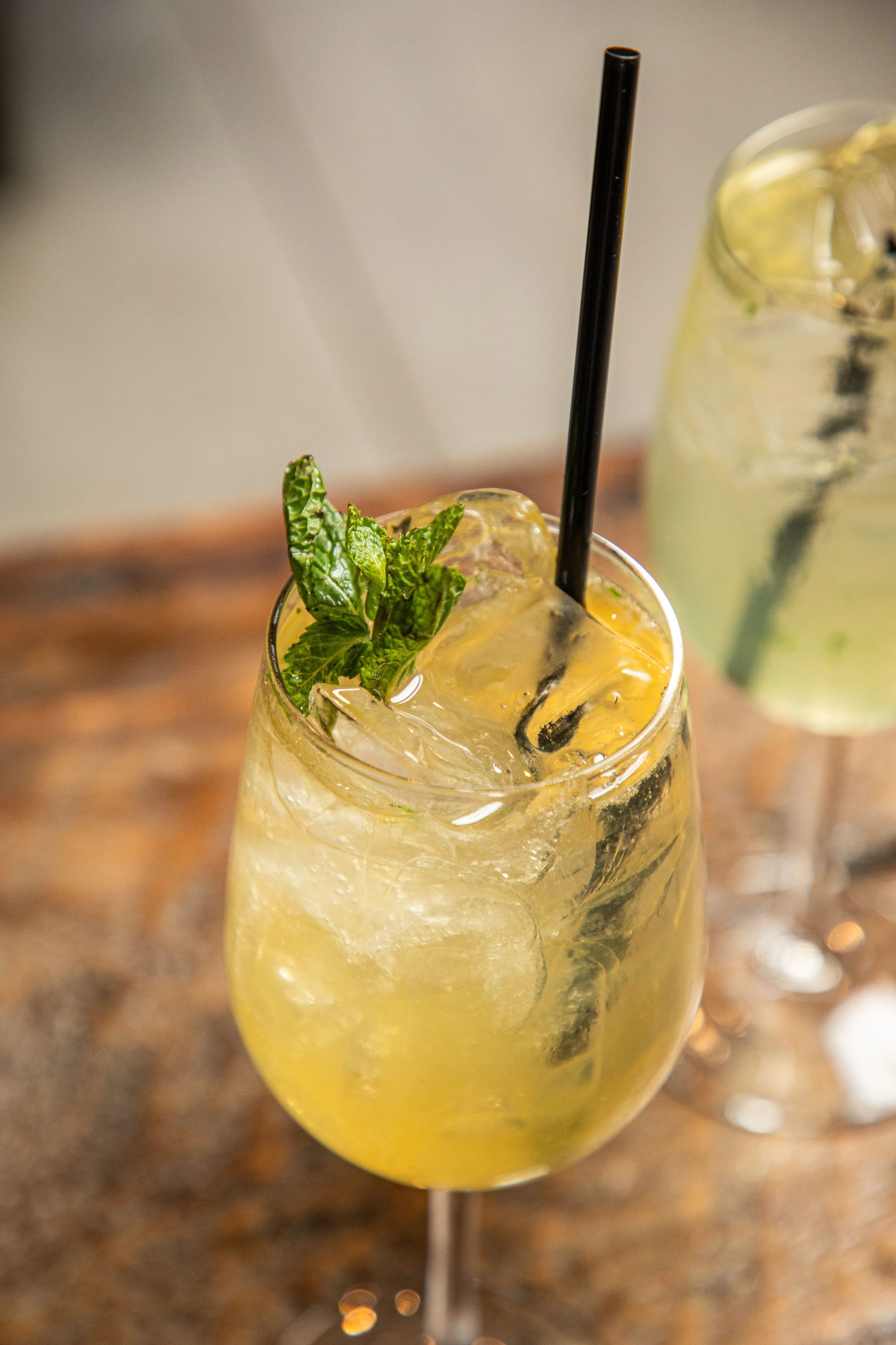 A glass of yellow-colored spritz cocktail with ice, mint leaves, and a black straw on a wooden surface with another similar drink blurred in the background.