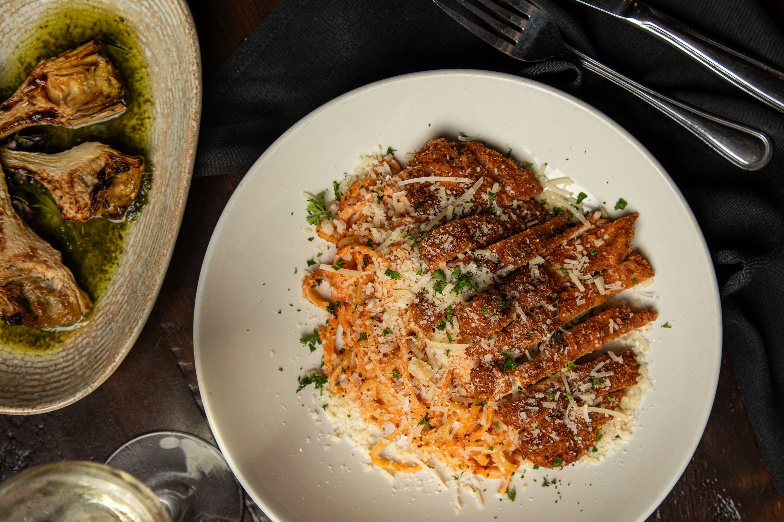 A white plate with breaded panne chicken, served with pasta and grated cheese, garnished with chopped parsley.
