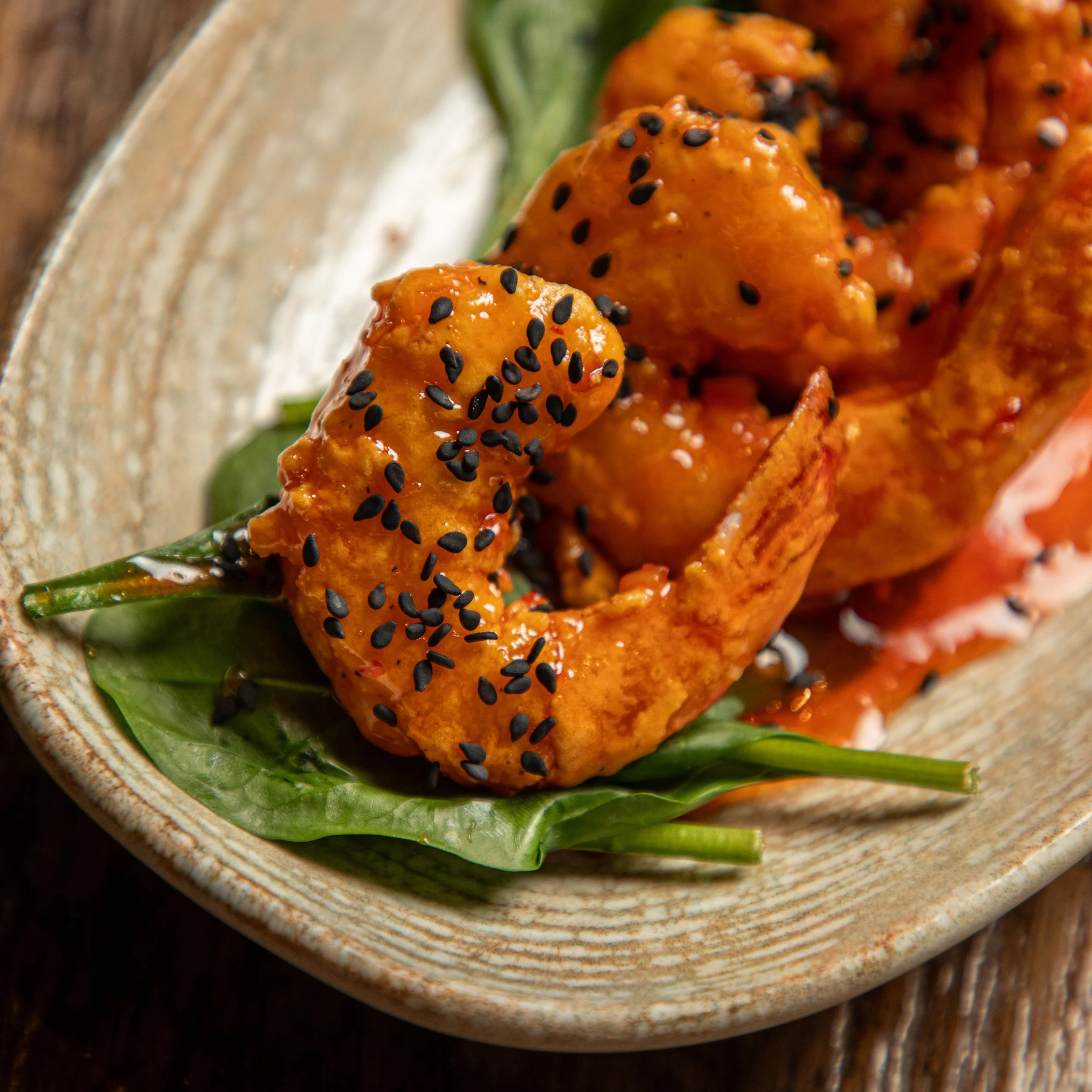 Close-up view of Bang Bang Shrimp coated in orange sauce and sprinkled with black sesame seeds, garnished with green leafy vegetables on a rustic ceramic plate.