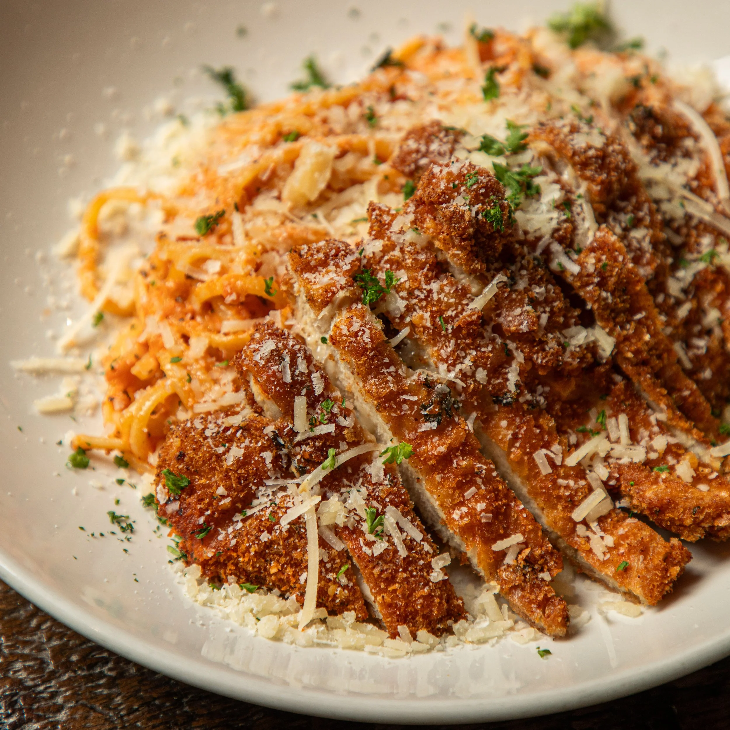 Close-up of a plate of fried chicken topped with grated cheese, chopped herbs, served with spaghetti in marinara sauce.