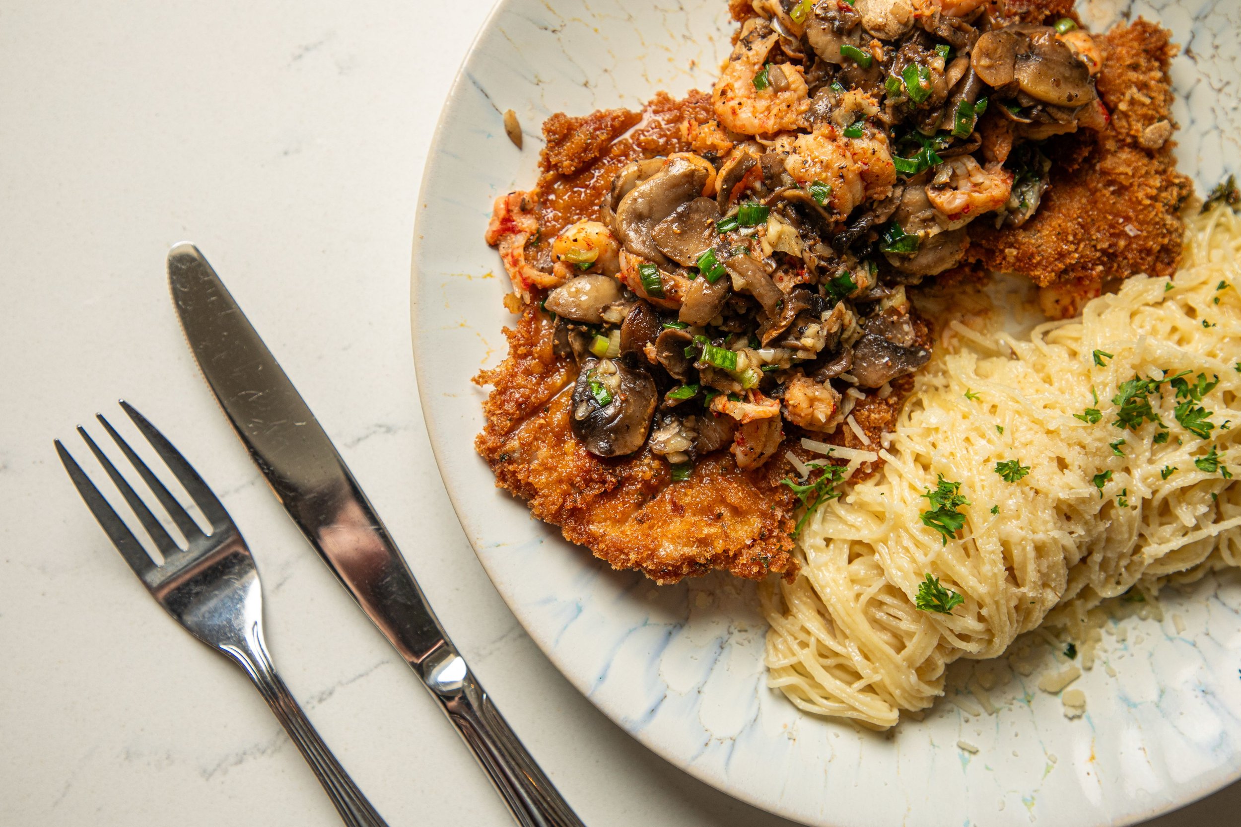 A plate of creamy pasta with parsley, breaded fried veal, and crawfish mushroom saute, set on a white surface with a knife and fork beside it.