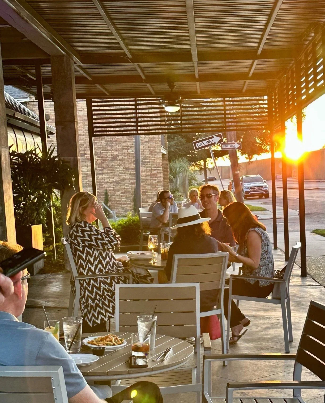 People enjoying dinner at an outdoor restaurant during sunset, with some seated at tables and others walking on the sidewalk. The setting features a shaded patio with wooden slats and hanging holiday lights.