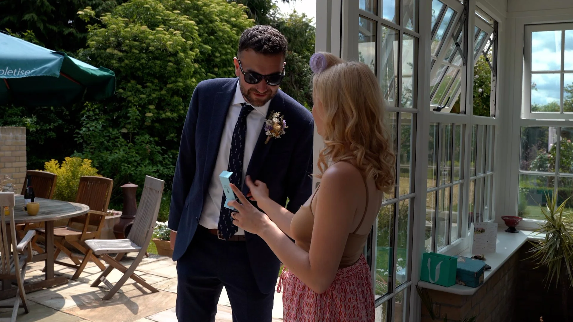 A man in a suit and sunglasses is looking at his phone while standing next to a woman with long curly hair, who is also focused on her phone, in a sunlit garden room with large windows and an outdoor patio with chairs and a table.