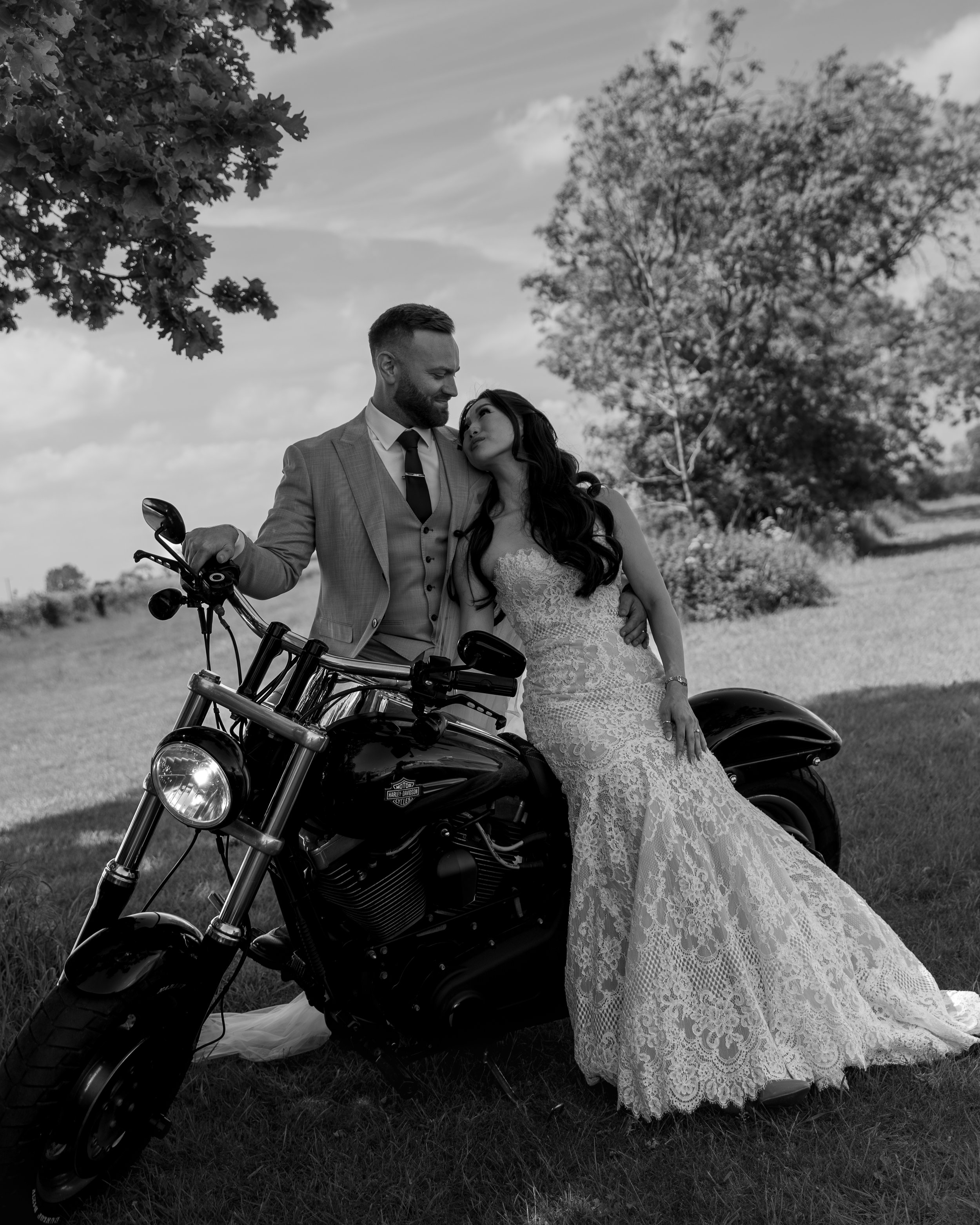 Black and white photo of a man in a suit and a woman in a wedding dress sharing a moment outdoors next to a motorcycle, with trees and a cloudy sky in the background.