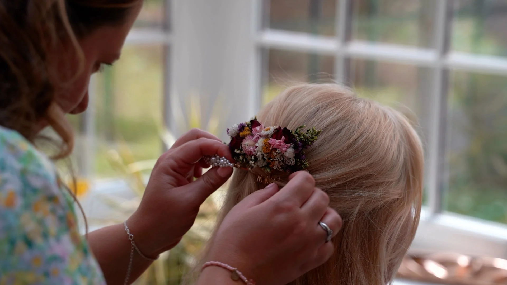 A woman attaches a floral hairpiece to a young girl's blonde hair inside a room with large windows and greenery outside.
