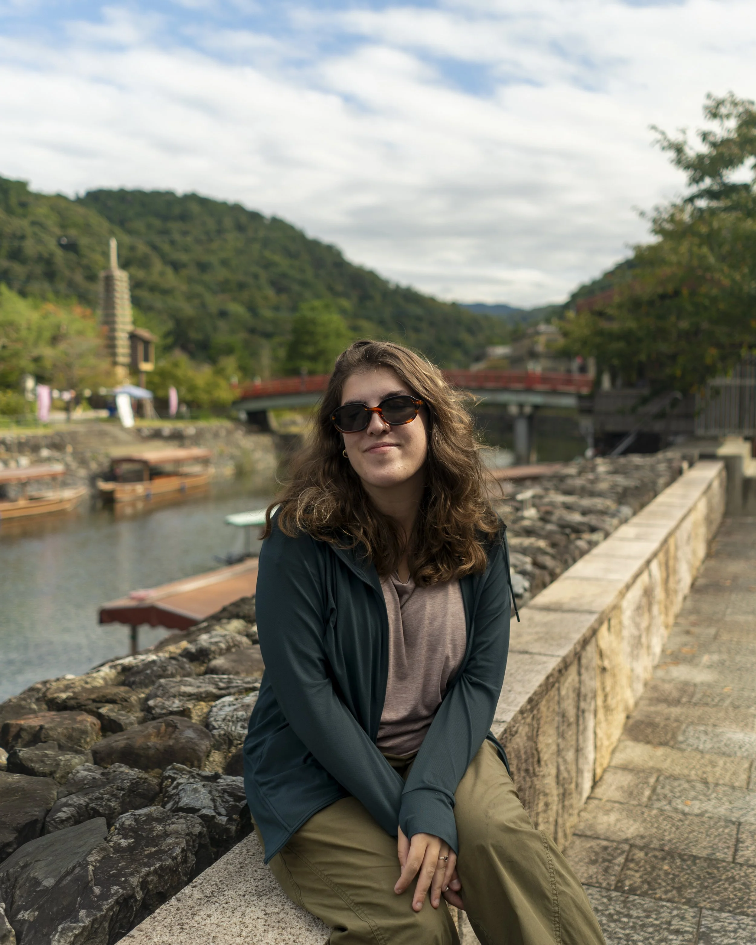 A woman with curly hair wearing sunglasses, a brown shirt, and a navy jacket, sitting by a river with a green, hilly landscape and a pagoda-like structure in the background, under a partly cloudy sky.