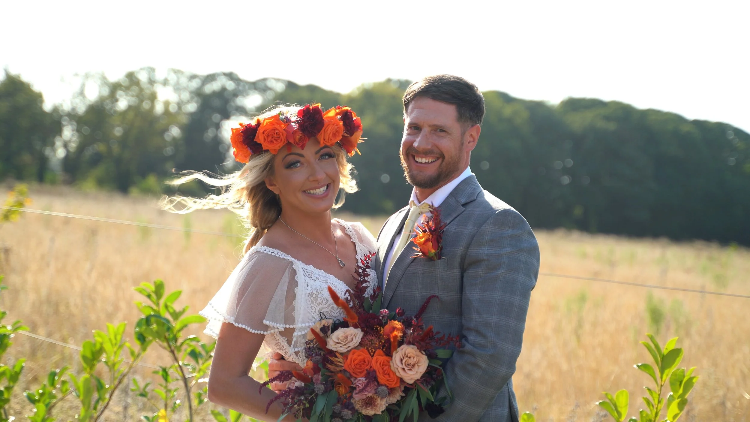 A smiling bride and groom standing in a field with trees in the background, the bride wearing a floral crown and holding a bouquet, and the groom in a suit with a boutonniere.