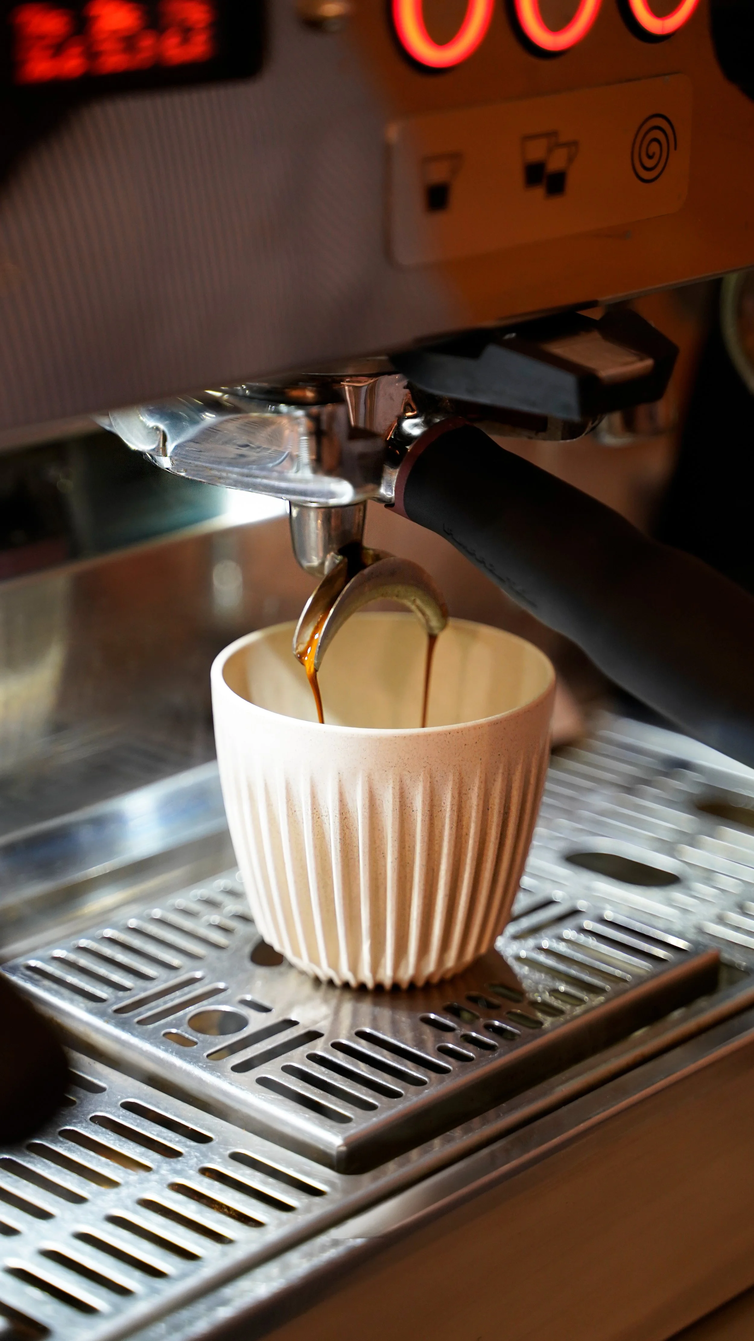 Close-up of a coffee machine dispensing espresso into a beige ceramic cup with vertical ridges, on a metal drip tray.