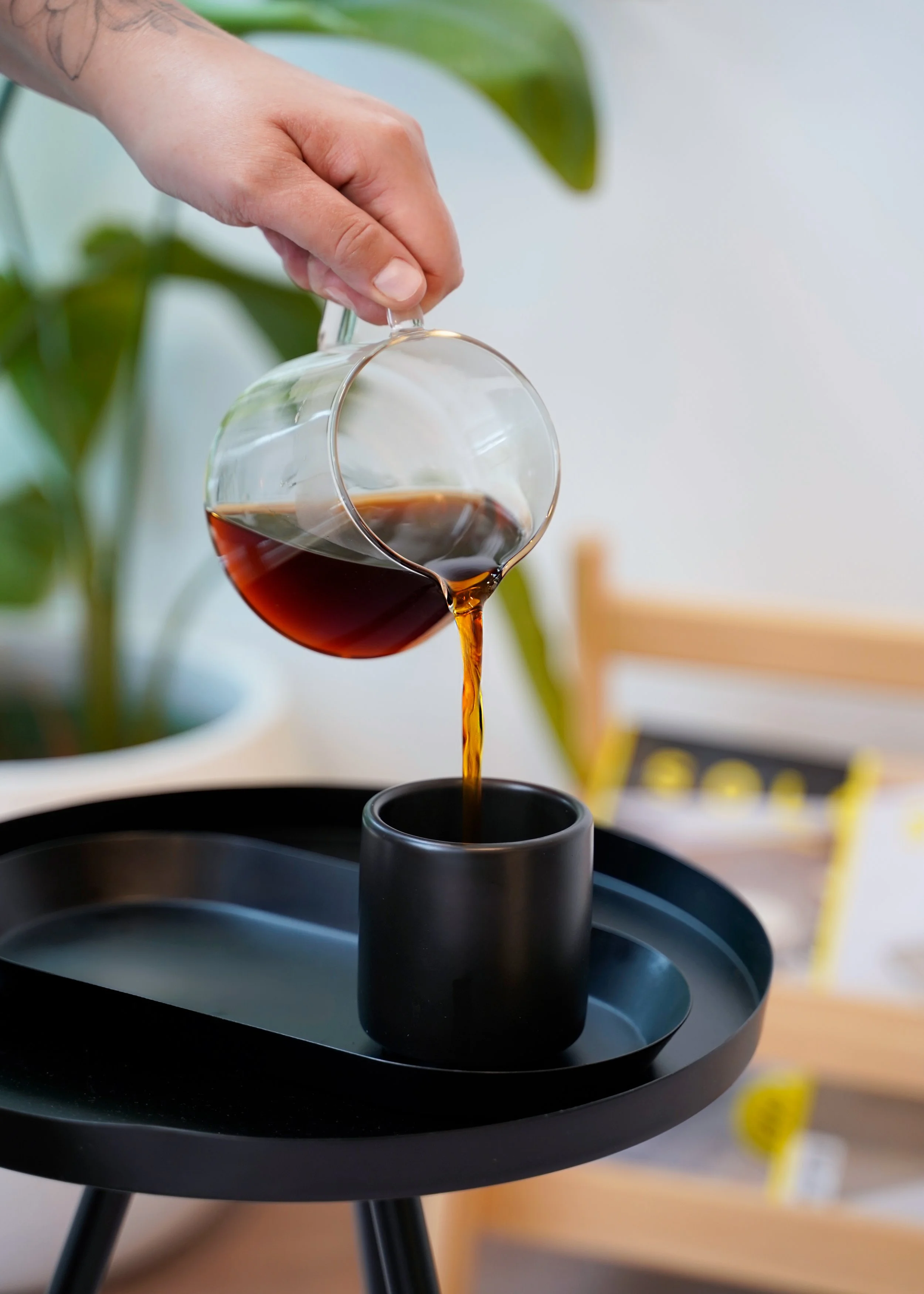 A person pouring dark coffee from a glass cup into a black mug on a black tray, with green plants and a blurred background.