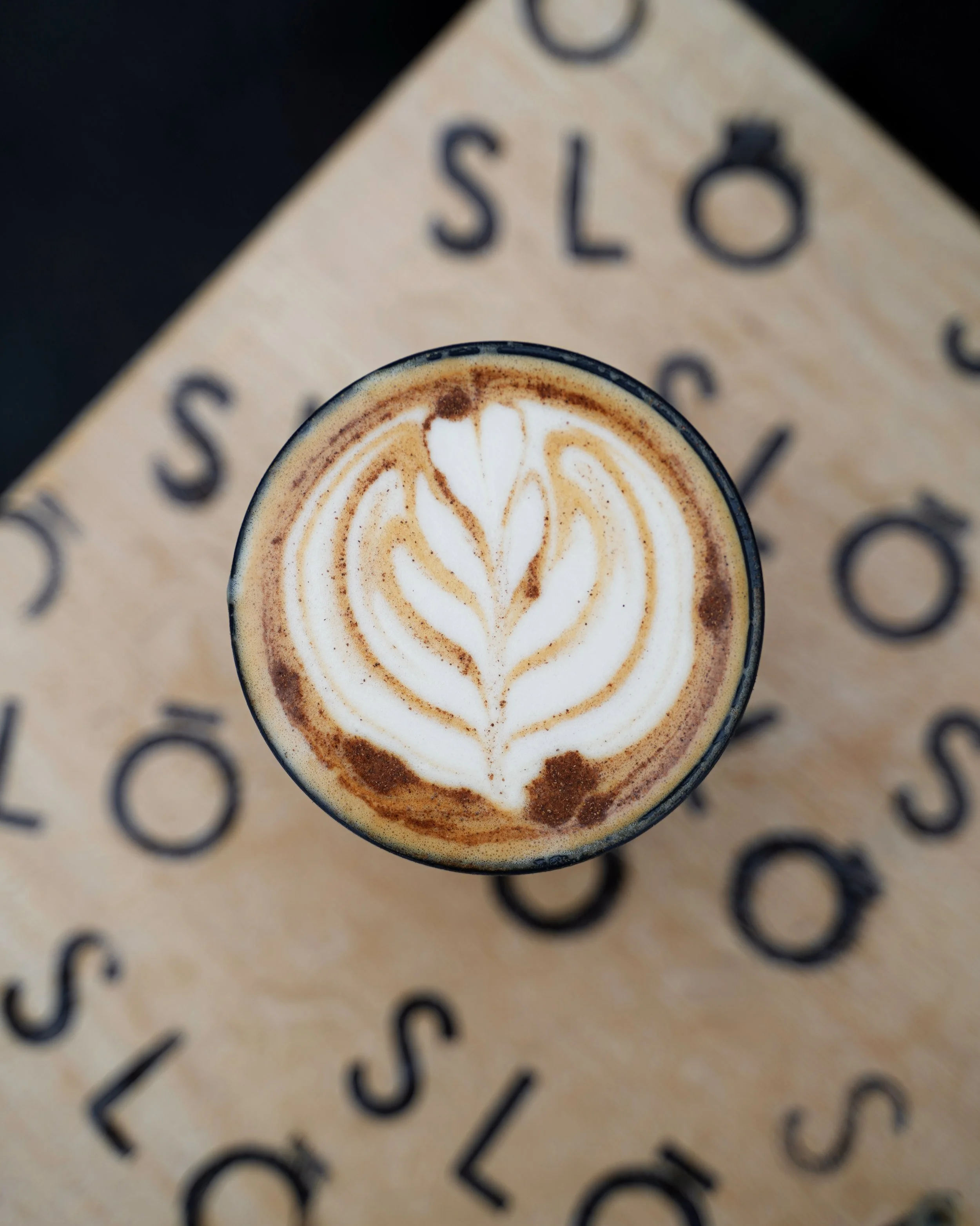 Top view of a latte with latte art in a black cup placed on a beige table with black printed text.