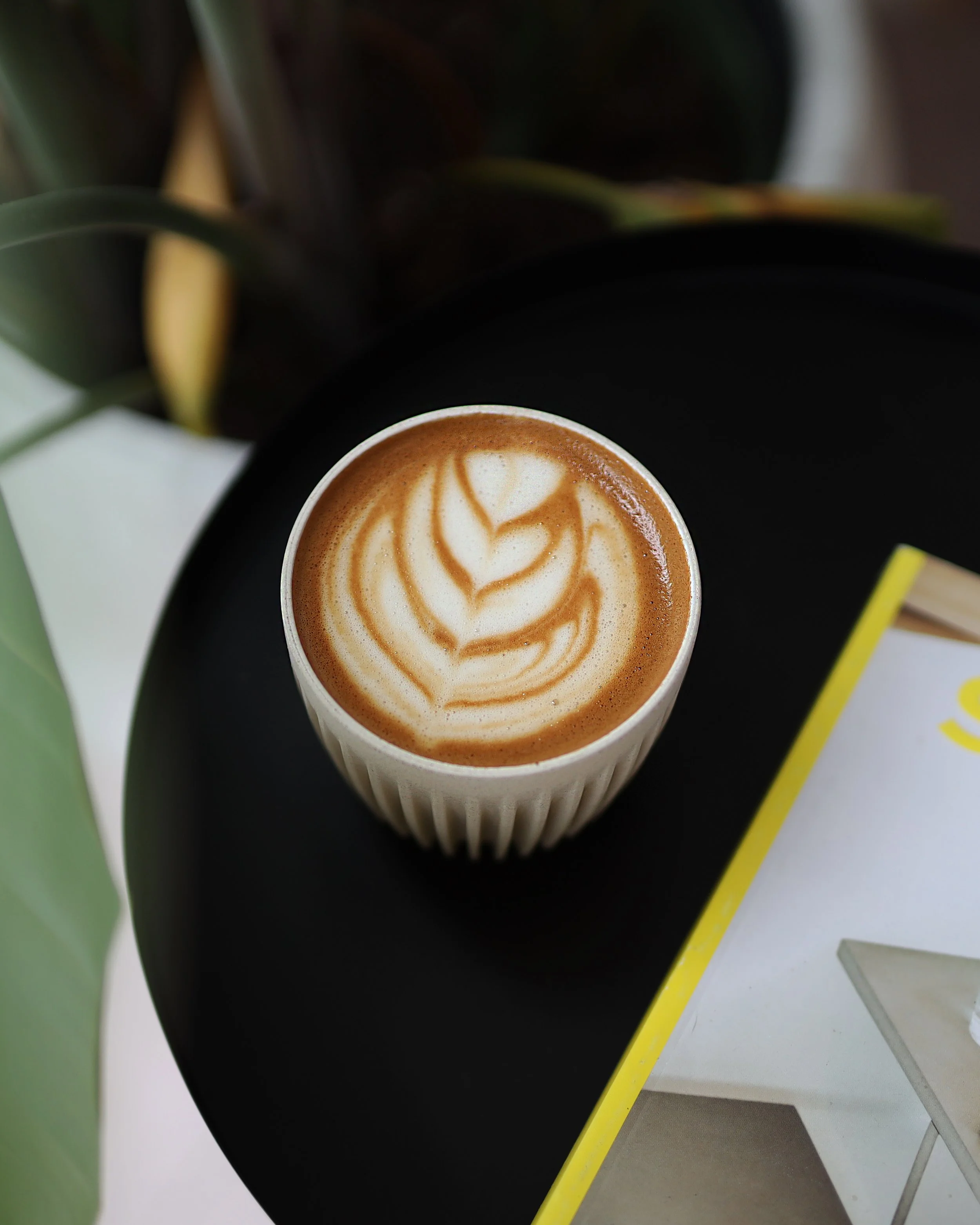 A cup of coffee with latte art on top, placed on a black surface, surrounded by various colored objects including a yellow straw, a green item, and white geometric shapes.