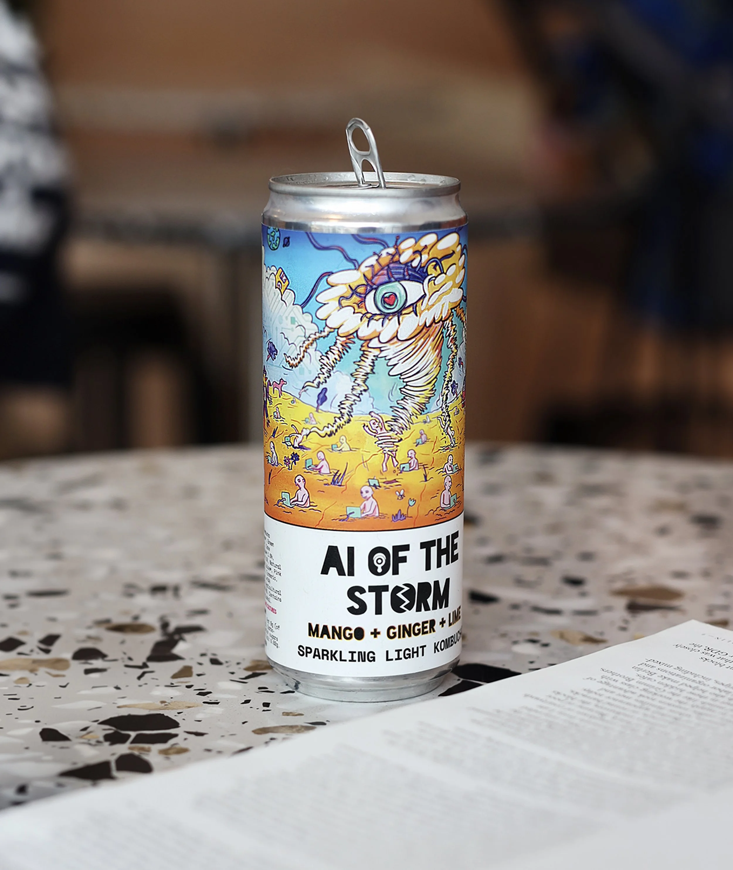 Can of Aloft the Storm sparkling light kombucha on a table with a patterned surface. The can features vibrant, colorful artwork with an eye and storm imagery. Part of a receipt or paper is visible at the bottom right corner.
