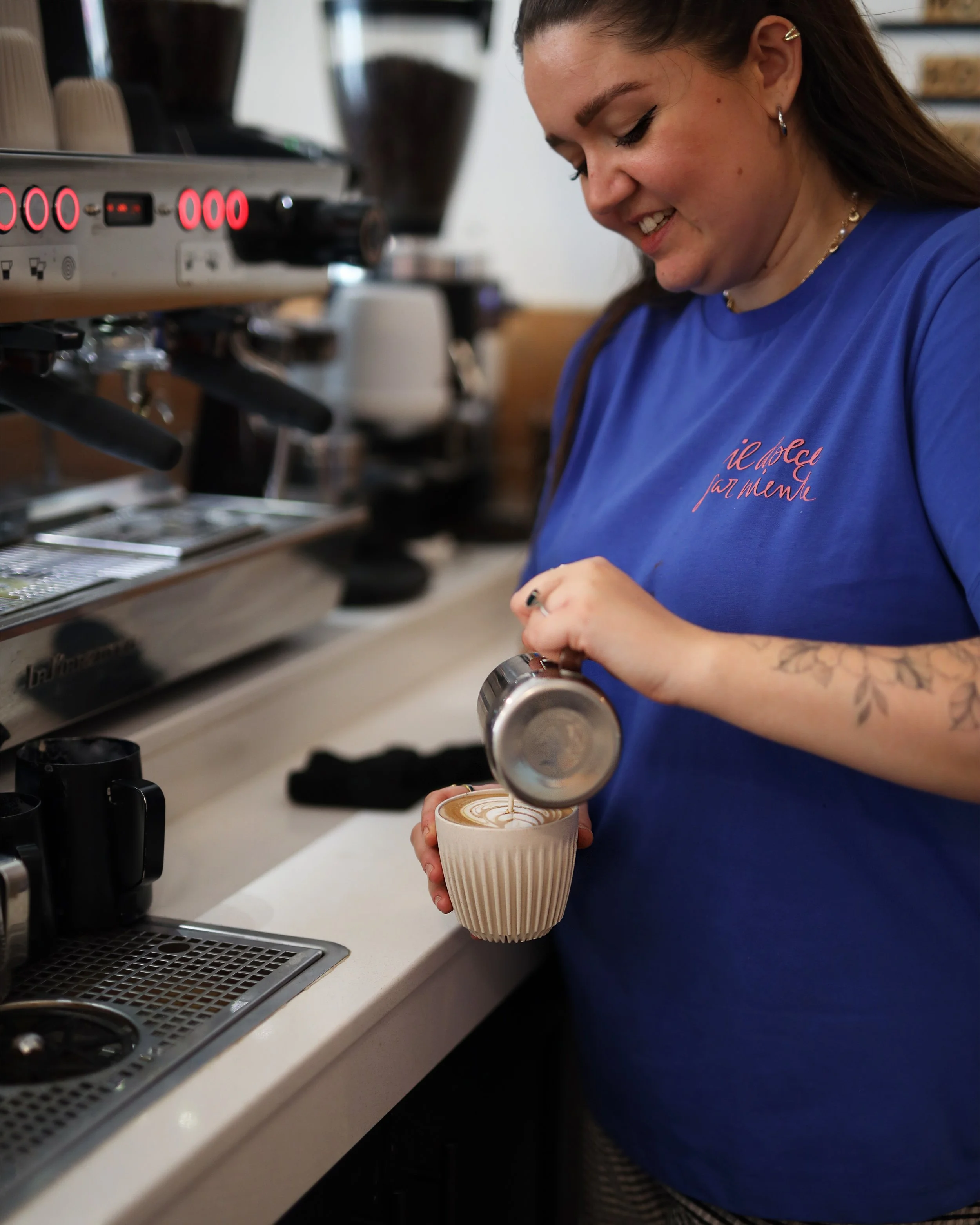A woman in a blue shirt preparing a latte with latte art in a coffee cup at a coffee shop.