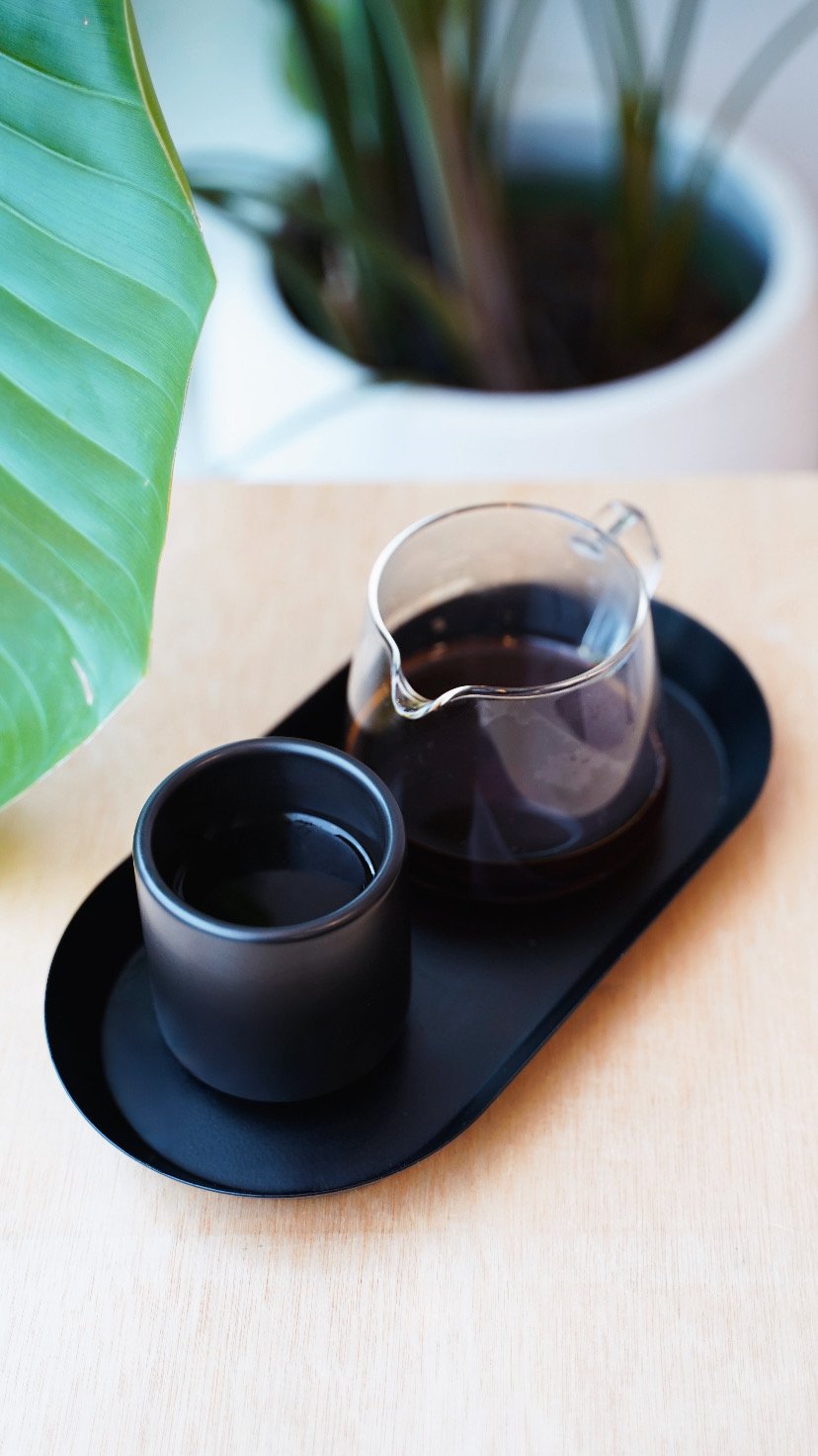 A black tray with a glass containing dark coffee and a small black cup, placed on a light wooden table. In the background, there's a large green plant with broad leaves.