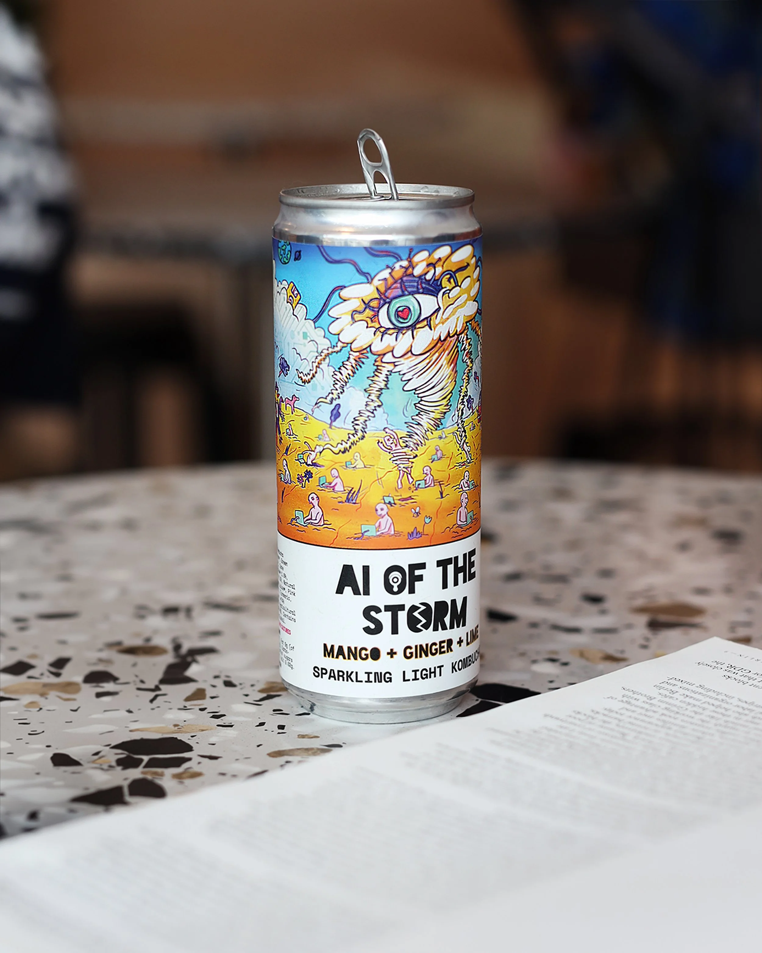 Aluminum can of Ai of the Storm sparkling light kombucha with colorful, artistic label featuring a large eye and yellow storm clouds, placed on a speckled surface with a piece of paper nearby.