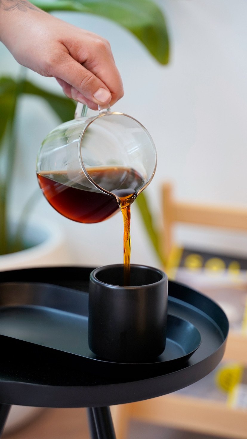 A hand pouring black coffee from a glass carafe into a black coffee mug on a black tray.