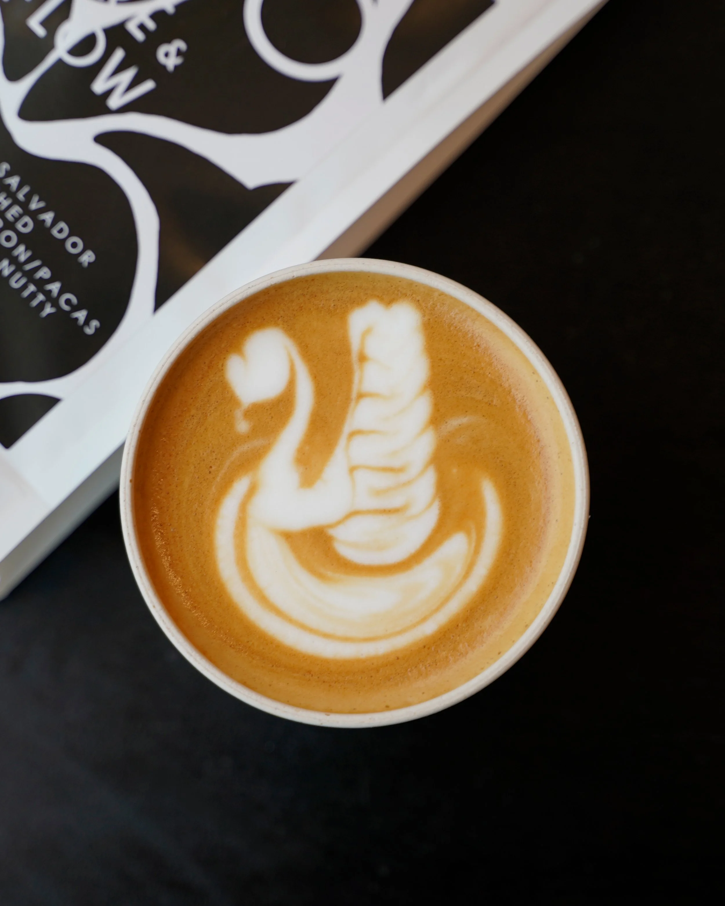 Close-up of a cup of coffee or latte with latte art on top, placed on a dark surface. Part of a white paper or menu with black printing is visible in the background.