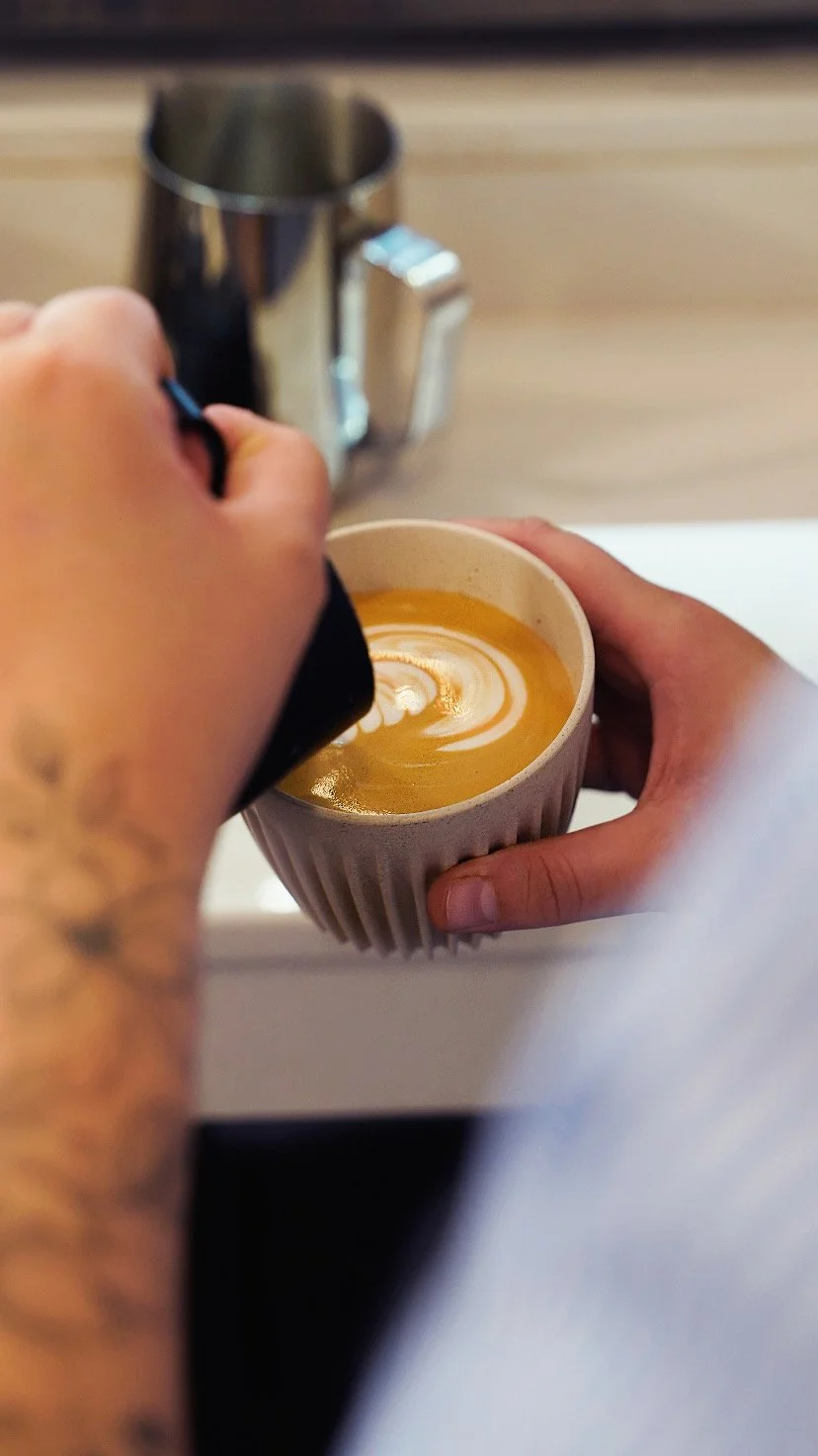 Person pours milk into a cup of coffee, creating latte art, with a silver milk frother in the background on a wooden surface.