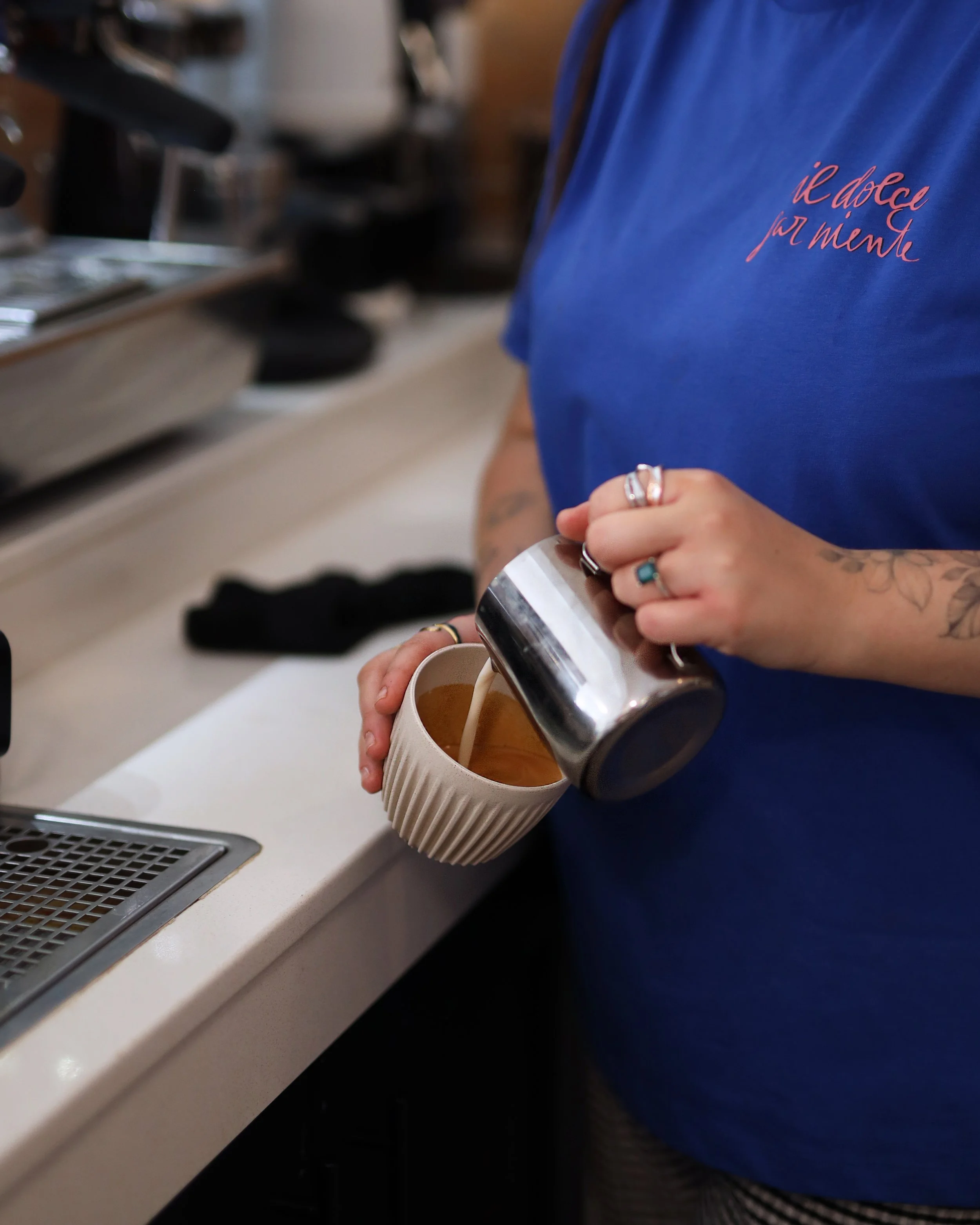 Person wearing a blue shirt standing at a counter and pouring a creamy beverage from a metal pitcher into a ridged ceramic mug.