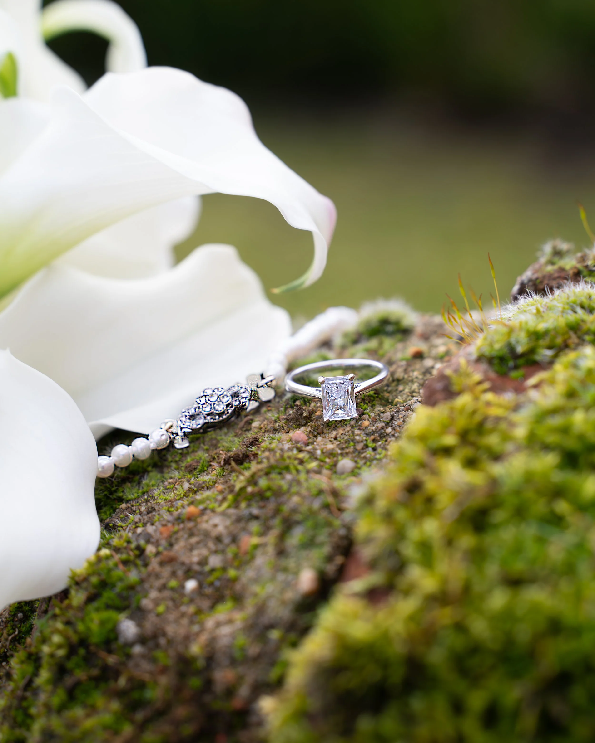 Close-up of a diamond engagement ring and wedding band on mossy ground with white flowers in the background.