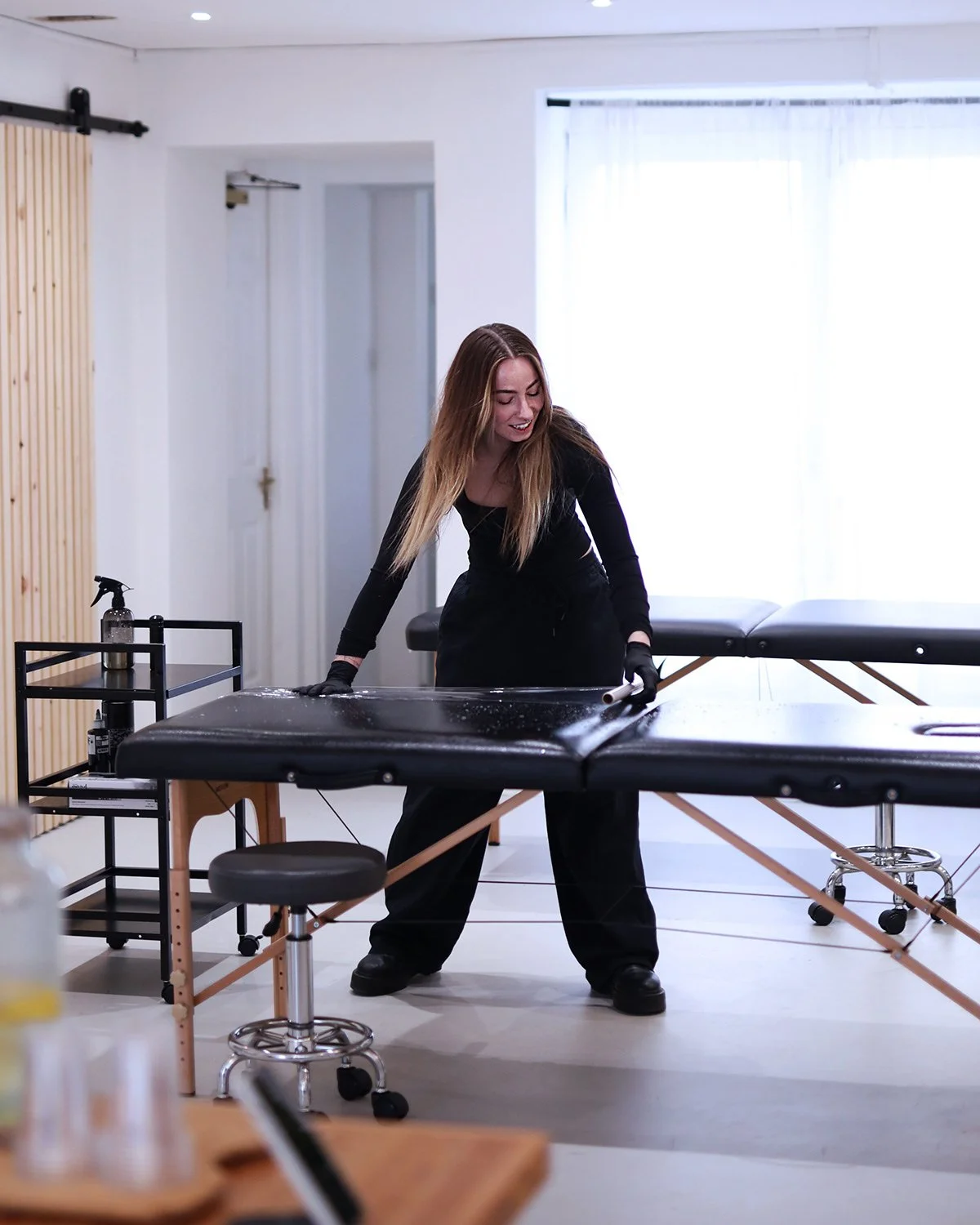 Woman cleaning massage table in a bright room with white walls and curtains, with a small cart holding cleaning supplies nearby.
