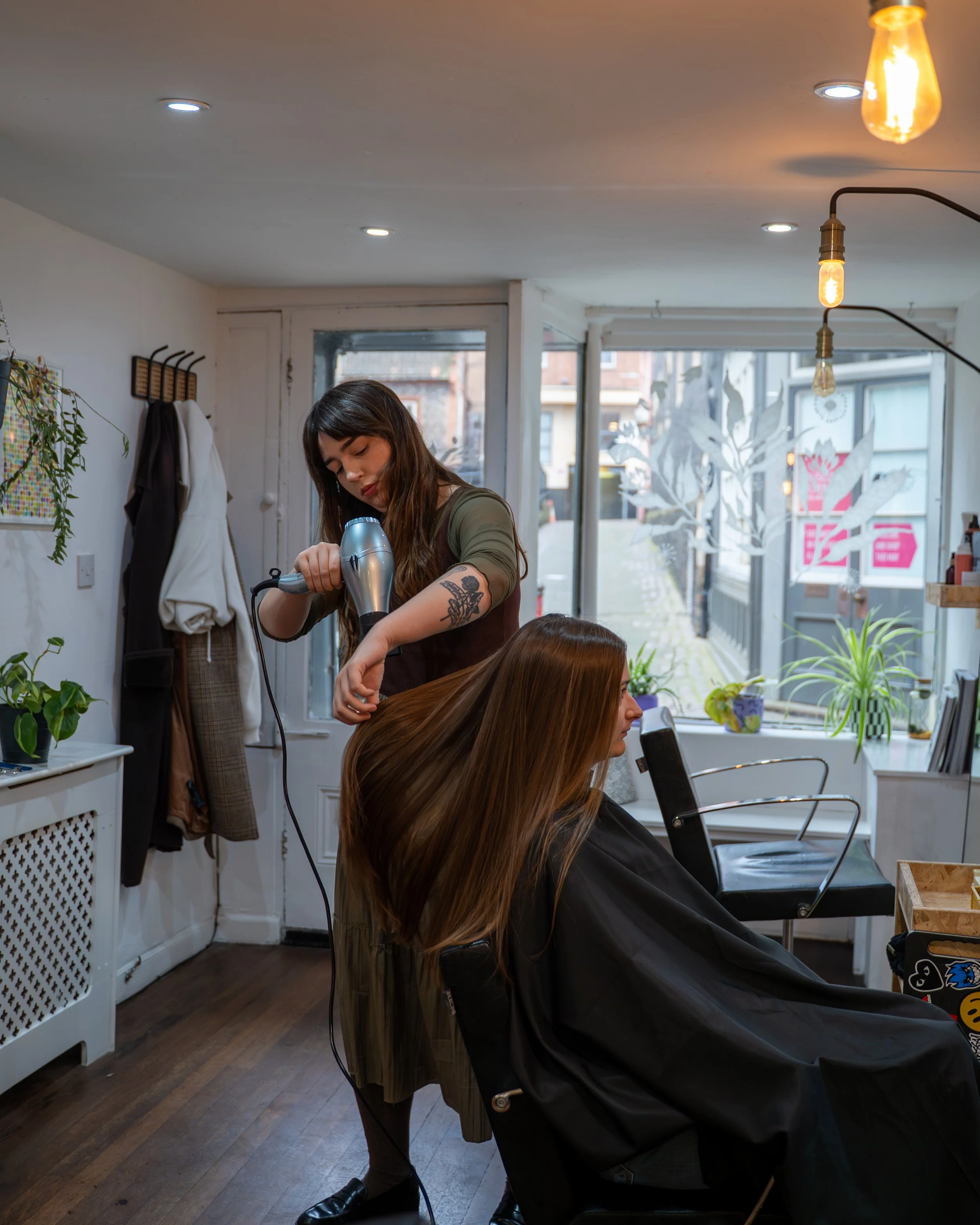 A woman with long brown hair and tattoos on her arm is getting her hair blow-dried by a hairstylist in a salon. The salon has a large window with plants and decorative paper cutouts outside, warm lighting, and a black seat for clients.