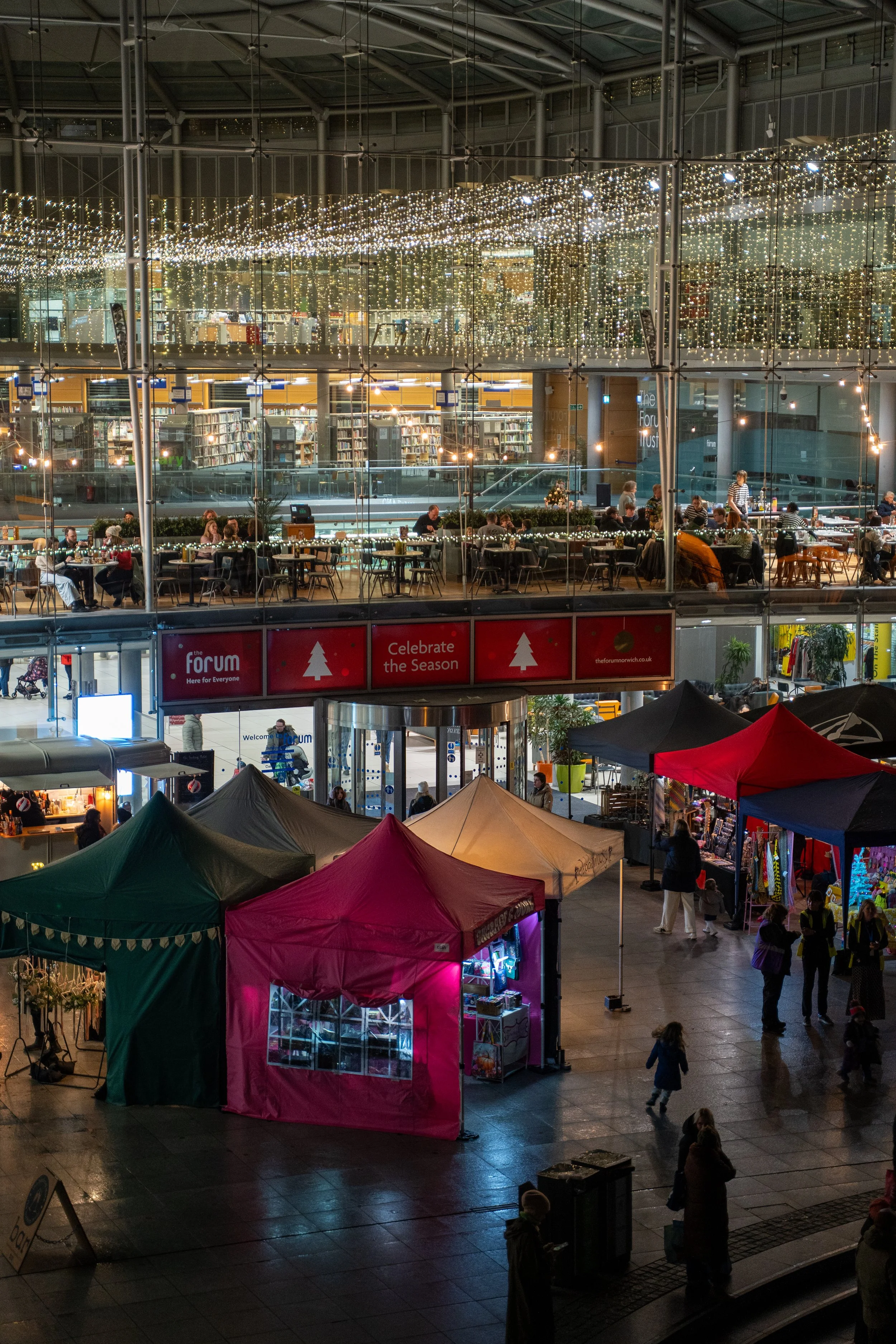 Night view of a multi-level indoor market with string lights decorating the ceiling. Several people are shopping and sitting at tables, with market stalls and tents set up on the ground level.