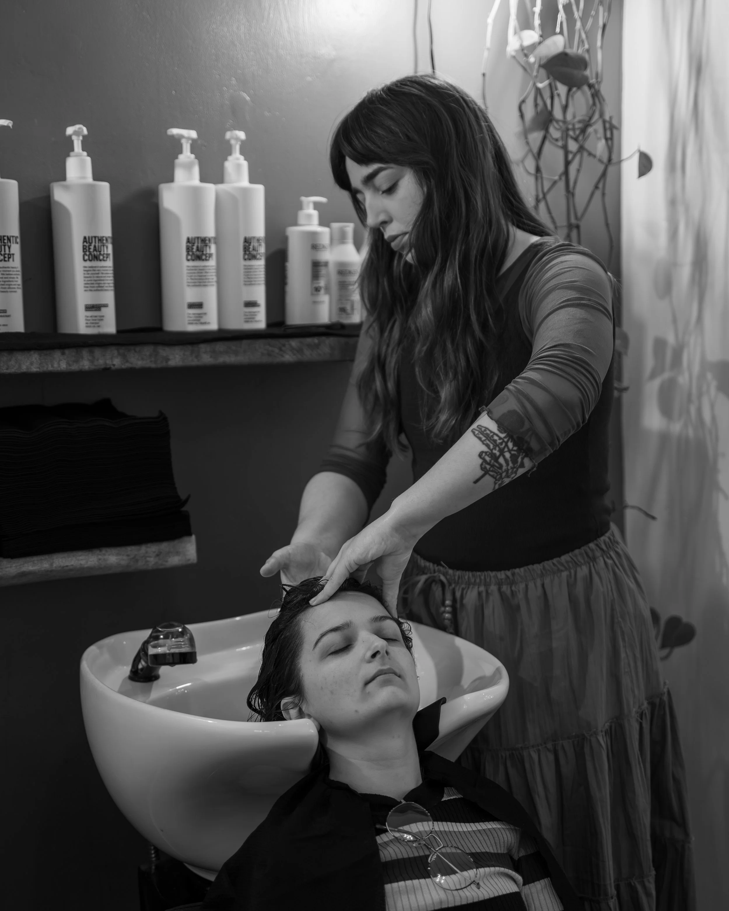 A woman wearing a dark top is getting her hair washed in a salon sink, with her eyes closed. A stylist with dark hair and tattooed arms is leaning over her, washing her hair. In the background, there are large bottles of hair products on a shelf and 