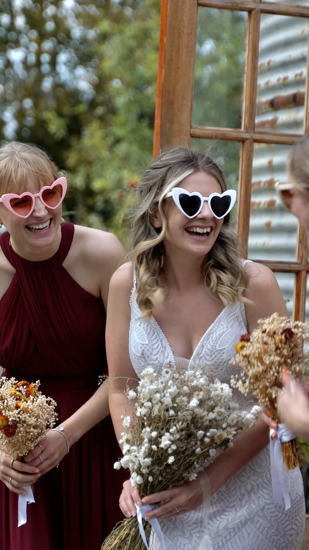 Two women wearing large heart-shaped sunglasses, smiling and holding bouquets of flowers, in an outdoor setting during daytime.
