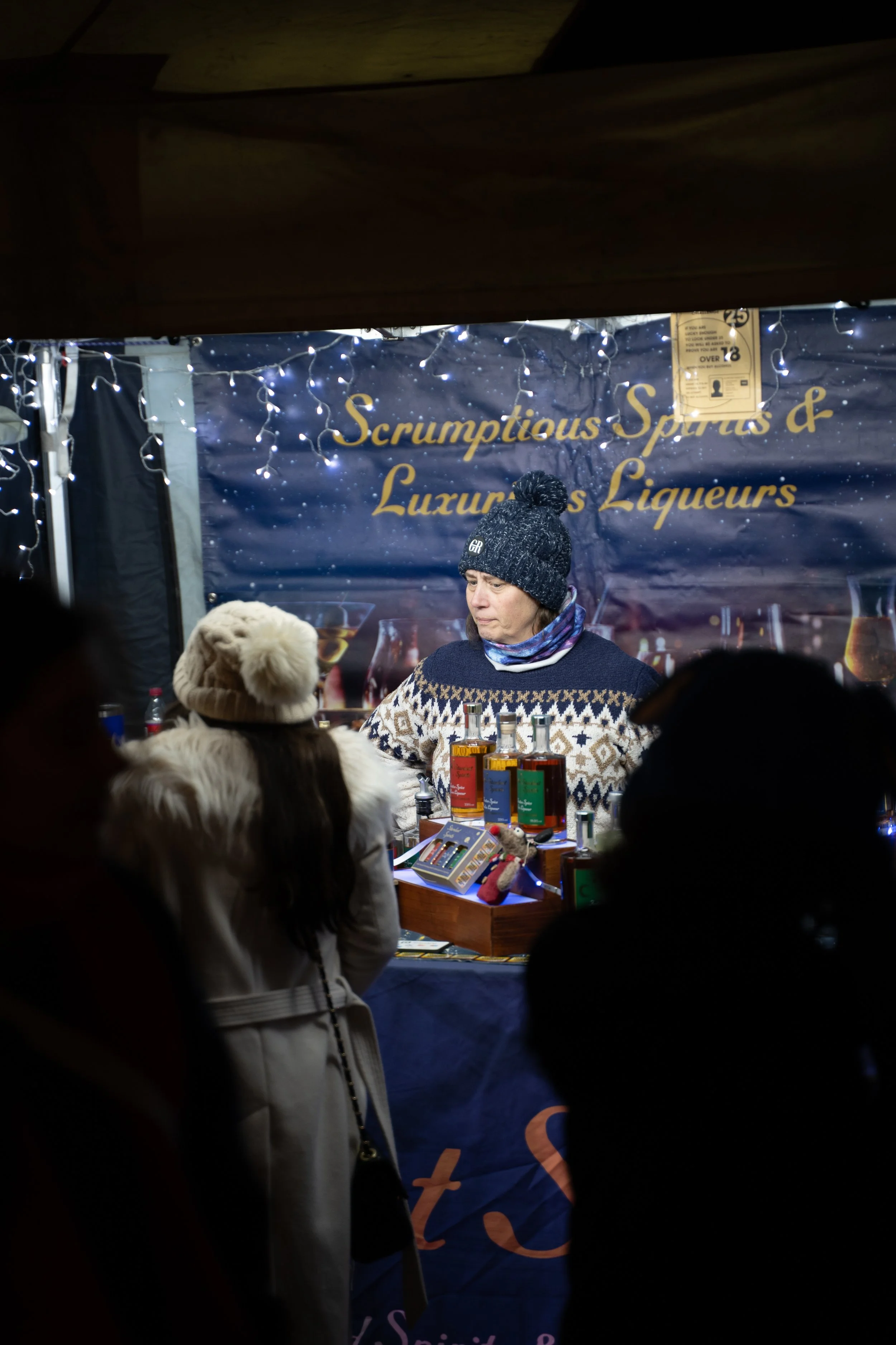 A woman wearing a black knit beanie and a patterned sweater stands in front of a booth with a dark blue banner that reads 'Scrumptious Spirits & Luxurious Liqueurs'. She is facing toward the right side of the image. There are bottles and items on a t