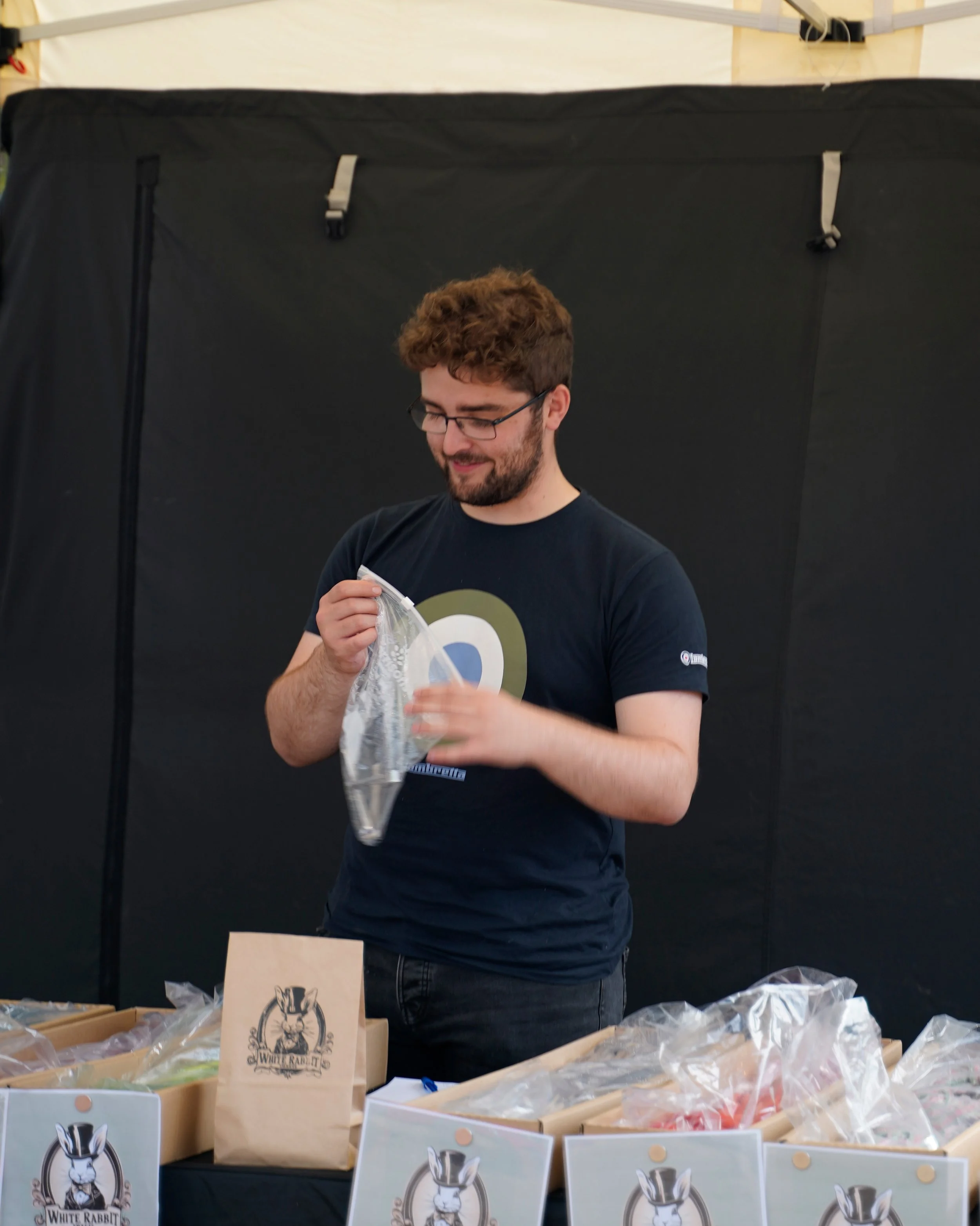 A man with glasses and a beard, smiling, standing behind a table with various packaged items and paper bags labeled 'White Rabbit,' at a market or event. The background is a black partition or wall.