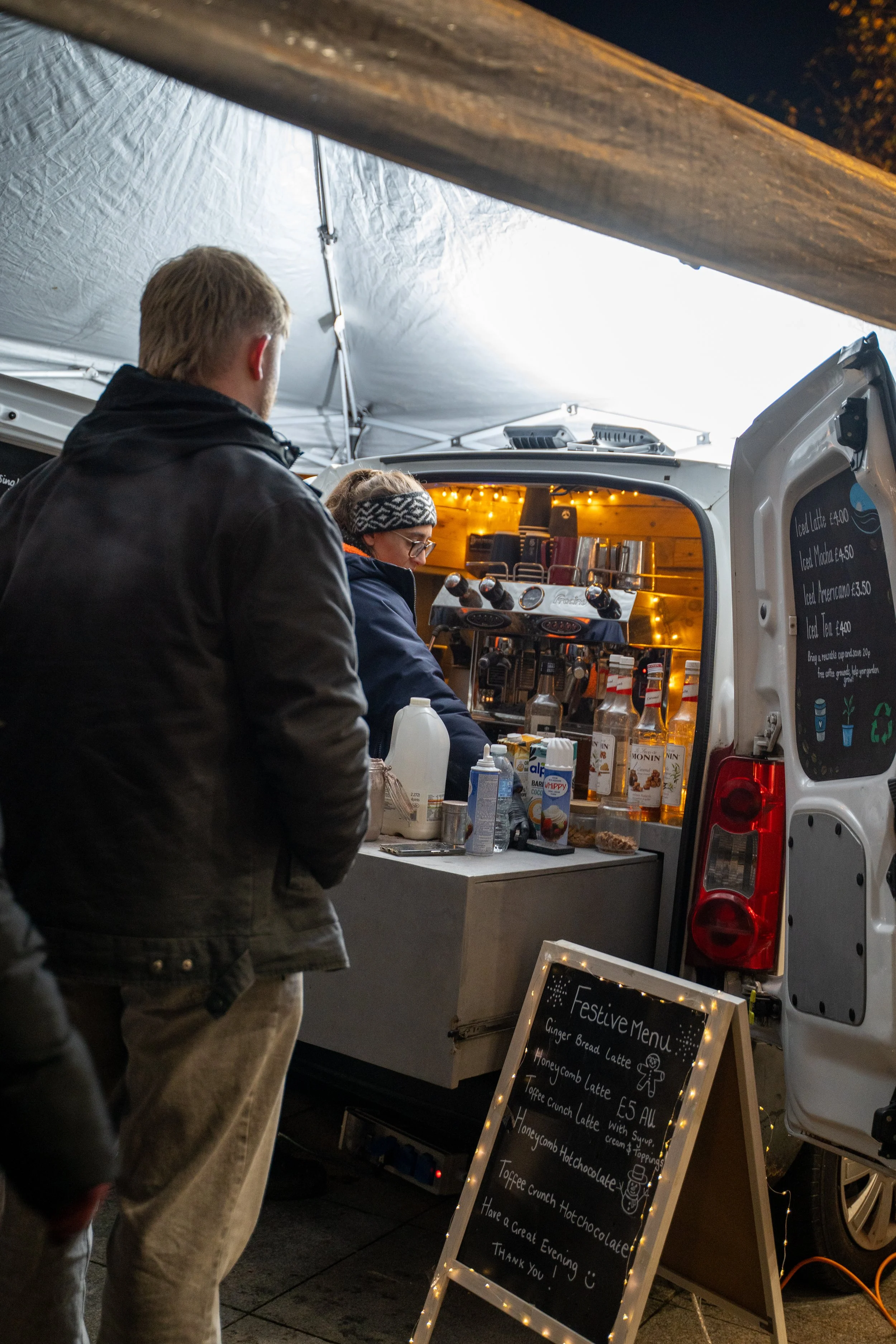 People at a Christmas market stand with a blackboard menu and drinks, outside at night.