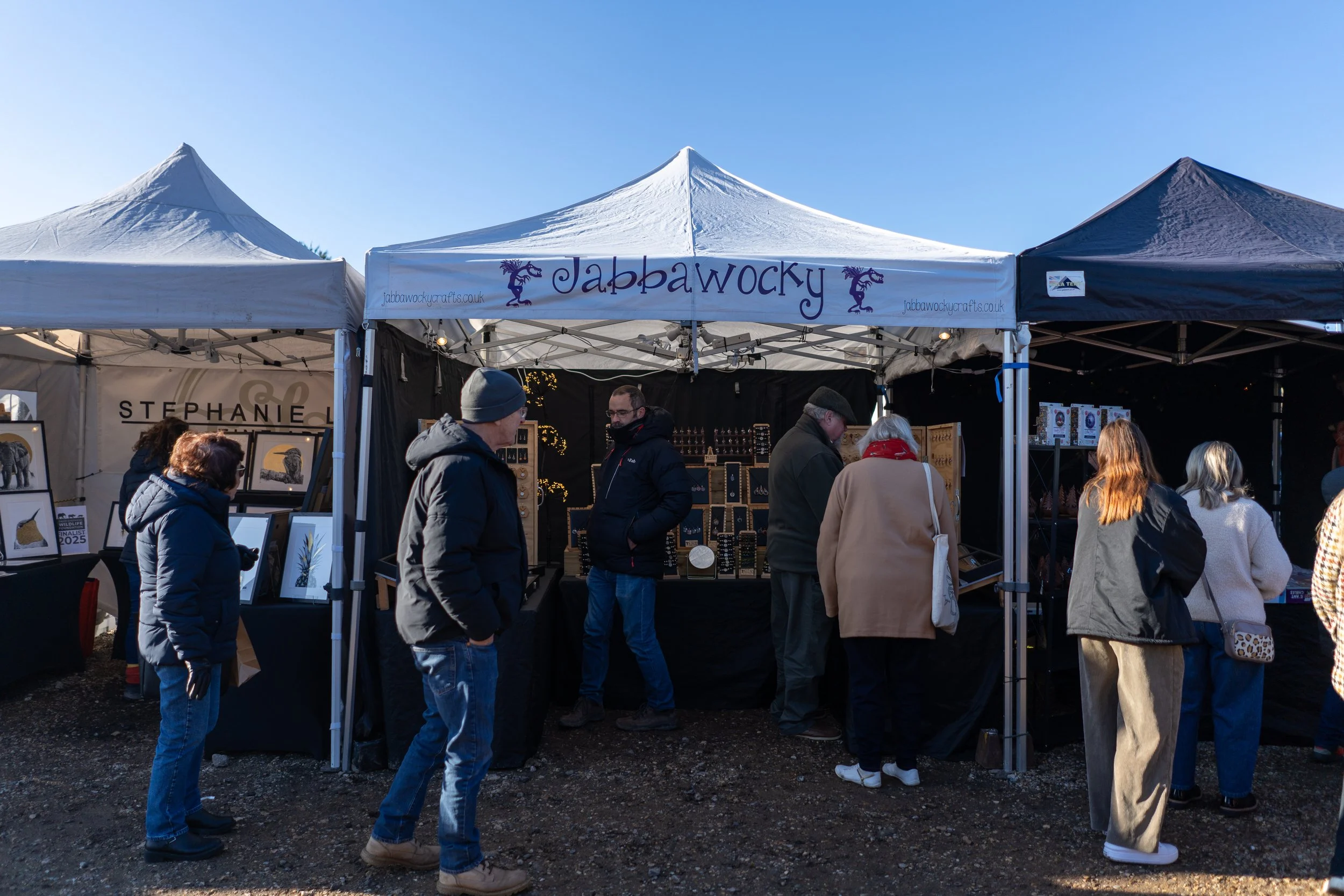 People browsing jewelry booths at an outdoor market with tents and a clear blue sky.