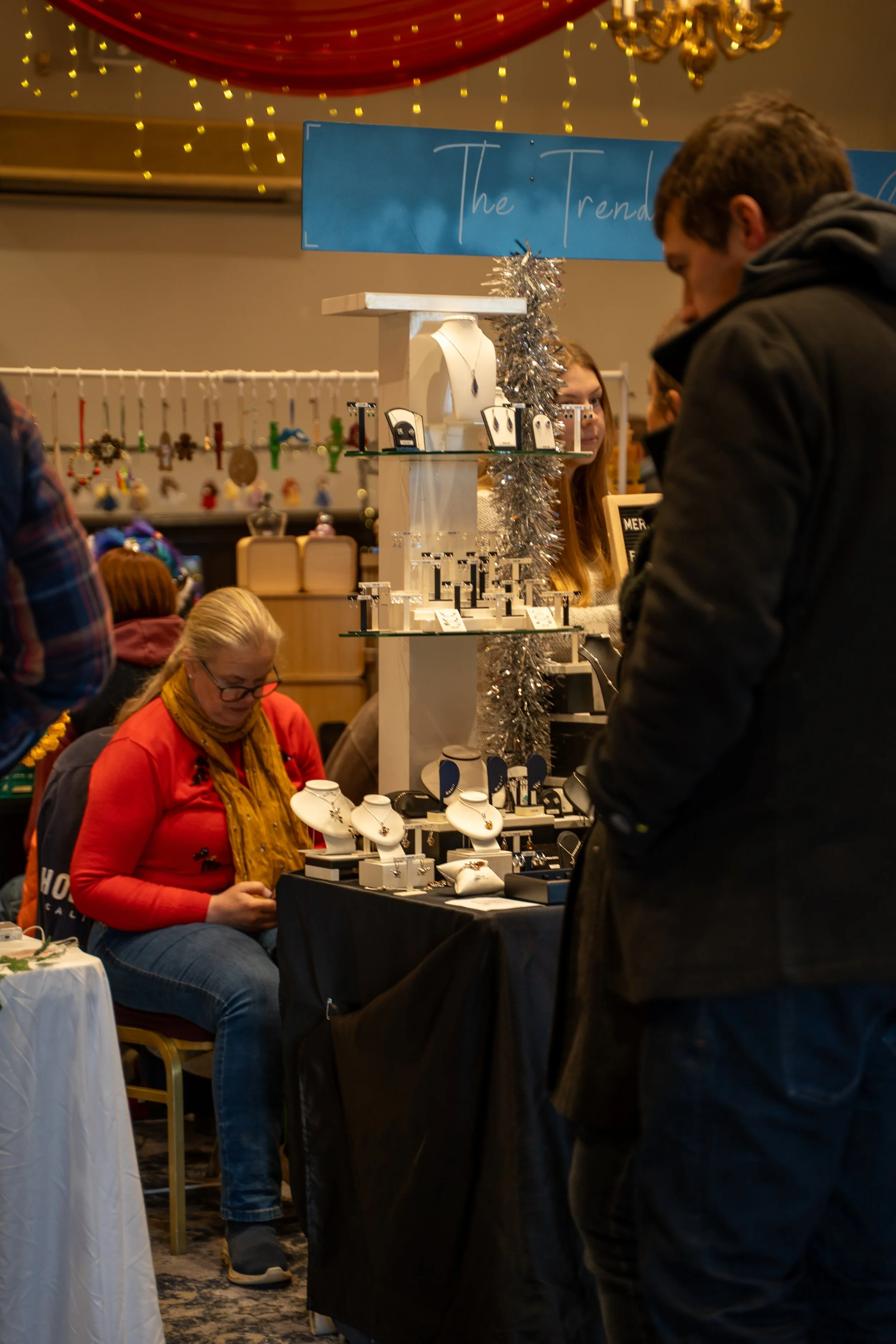 Jewelry vendor at a market stall with necklaces and bracelets, with customers browsing and a sign that reads 'The Trends' in the background.