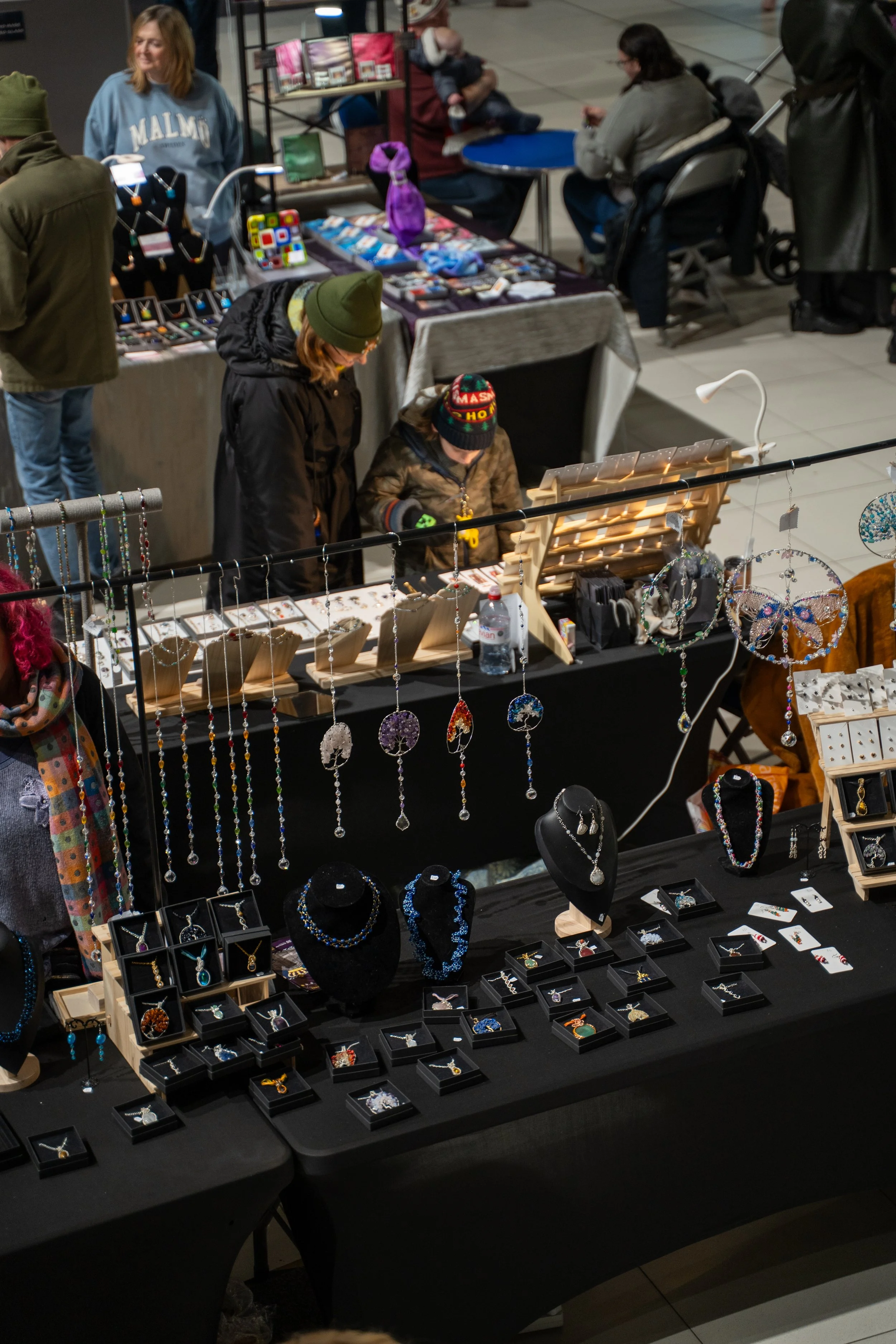Jewelry display booths at a craft fair with necklaces, rings, and earrings, with people browsing and shopping.