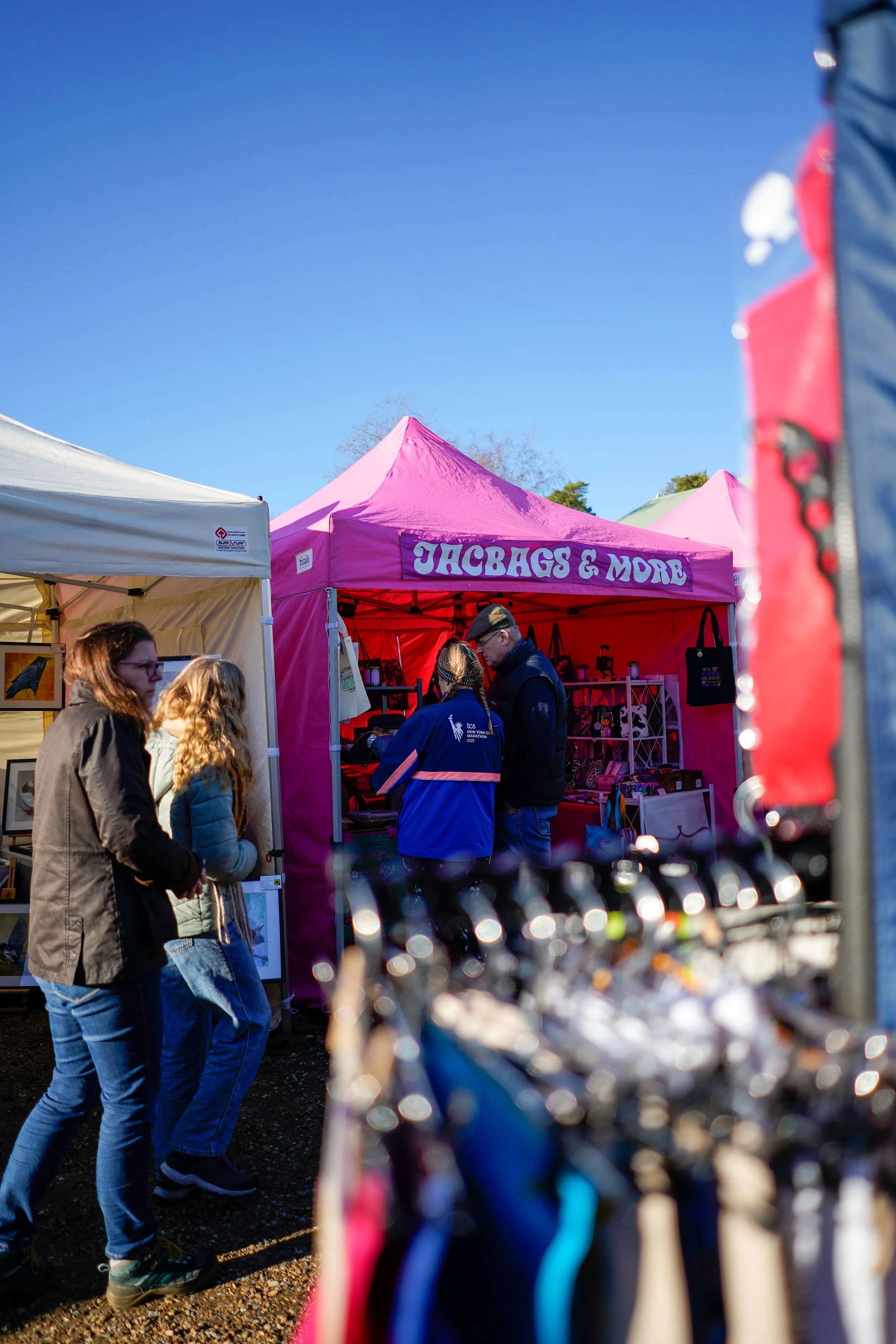 People shopping at an outdoor market with colorful tents, including a pink tent labeled 'JACKBAGS & MORE,' under a bright blue sky.