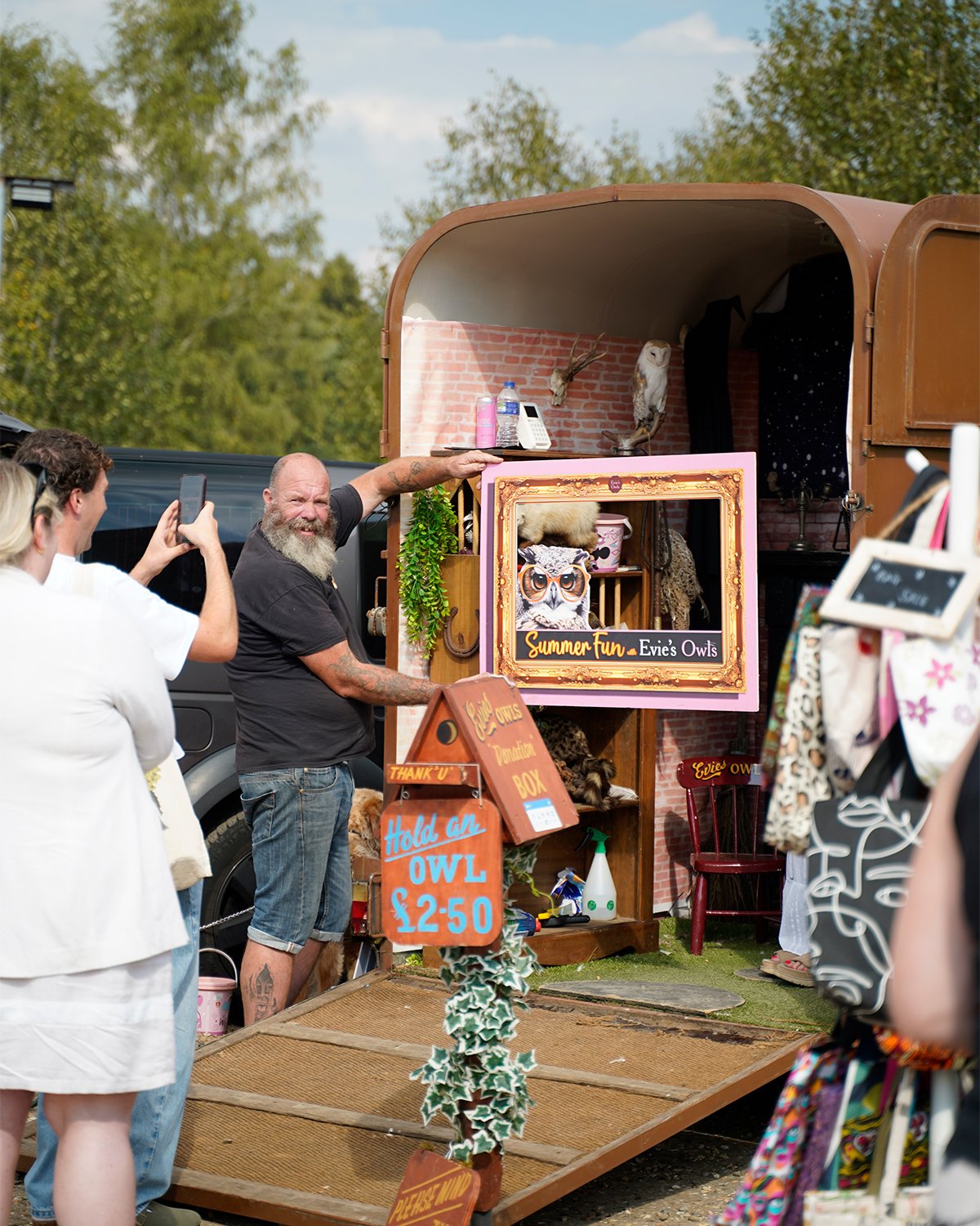 A man with a gray beard and tattoos on his arms is smiling and posing next to a mobile Owl exhibit called 'Summer Fun - Evie's Owls.' Several people are taking photos of him. The exhibit features signs with prices to hold owls and decorated owl-themed displays.