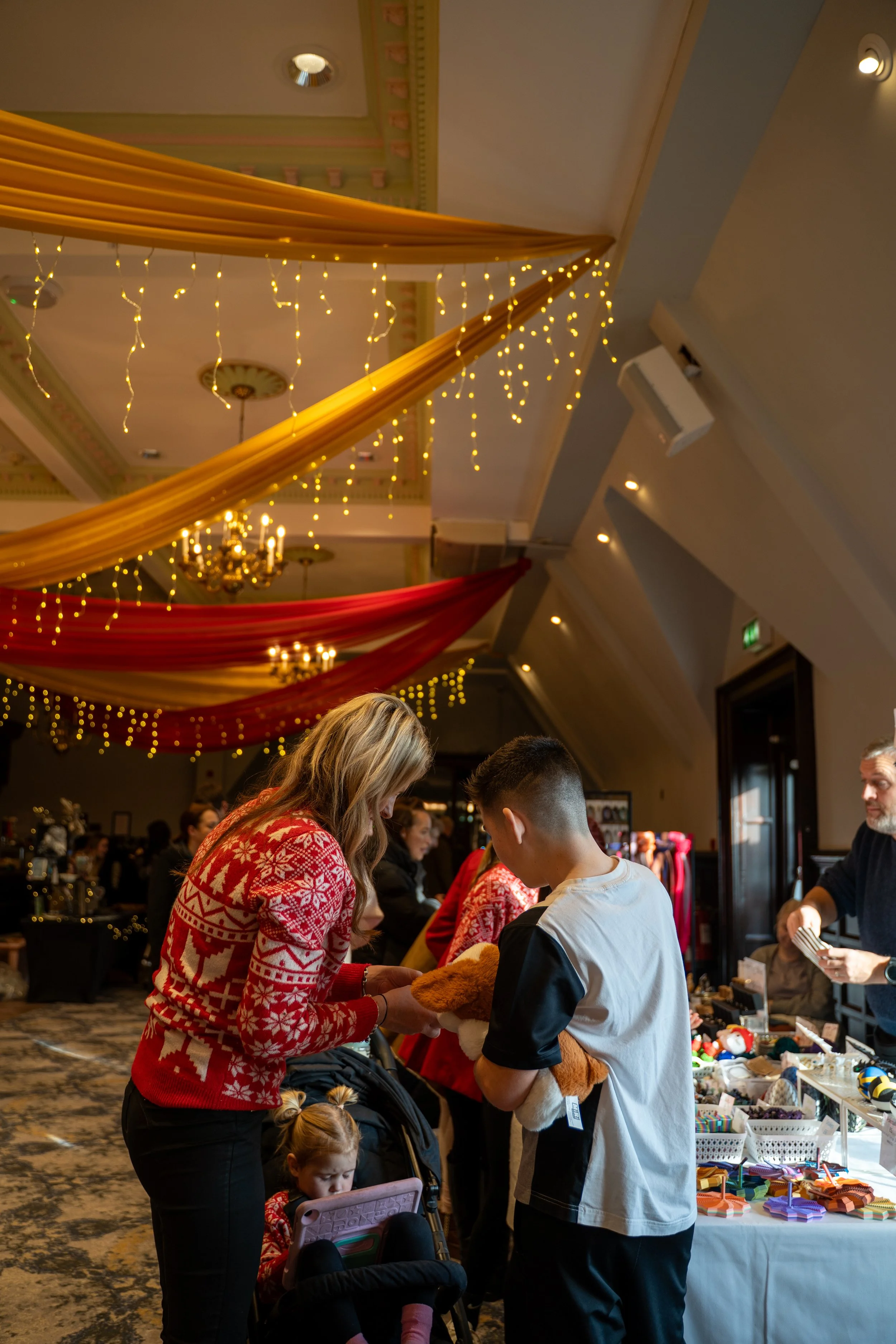 People shopping at a Christmas market stall with holiday decorations, including fairy lights and red and gold drapes, inside a decorated ballroom or event hall.