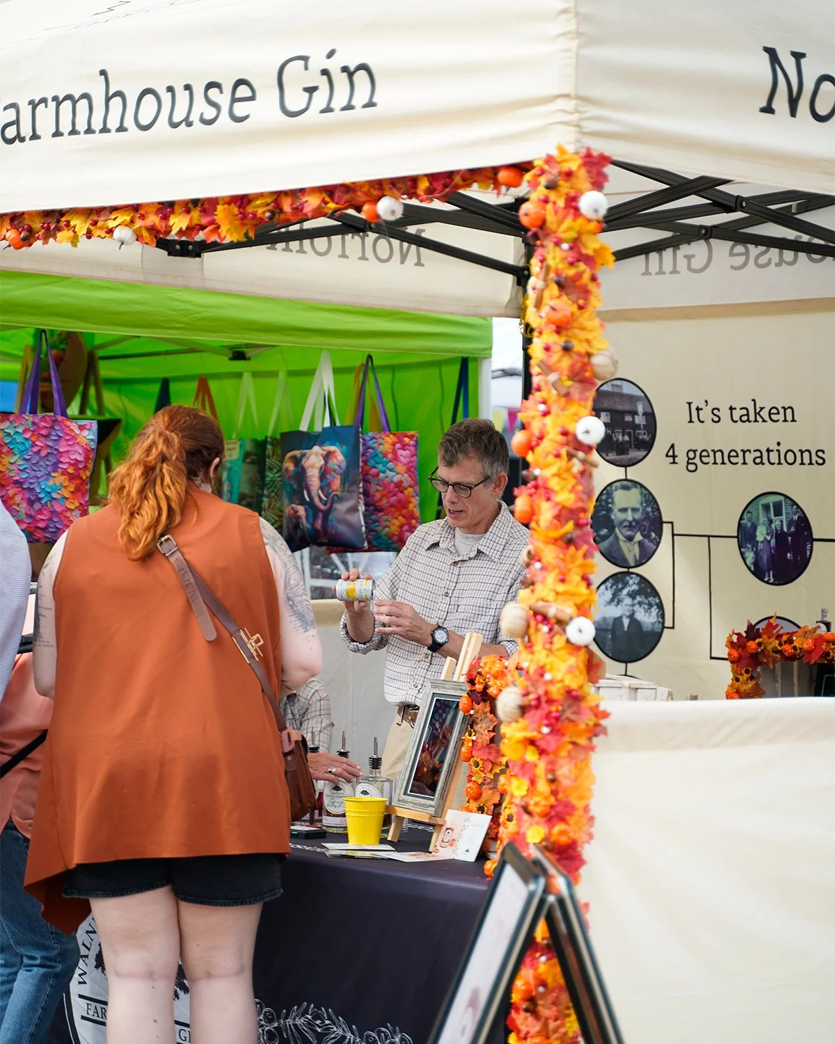 A man and a woman talking at a booth decorated with orange and yellow fall leaves and flowers, with colorful tote bags hanging behind them, at an outdoor market or fair.