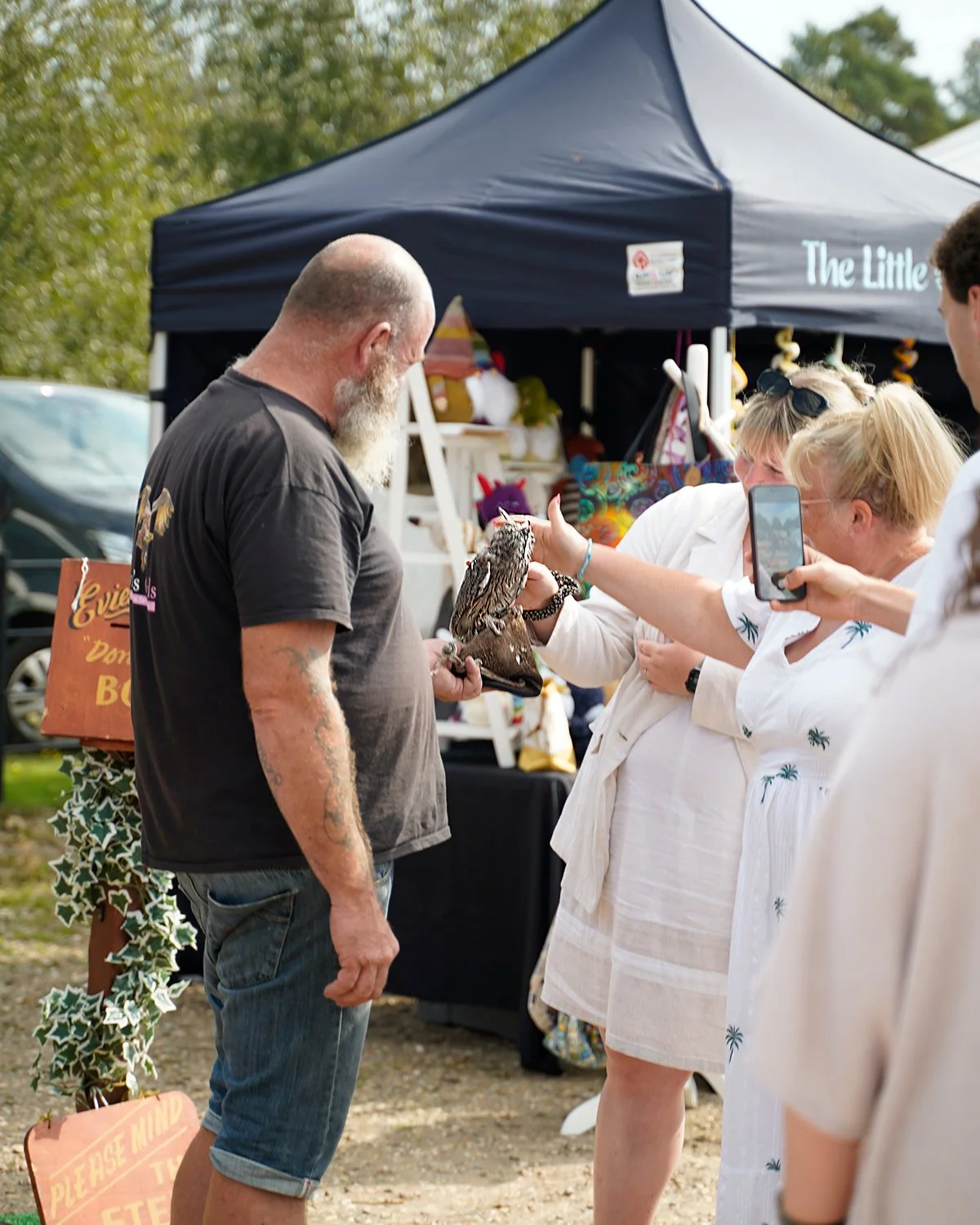 A woman is holding a small owl on her hand while a man looks at it. The woman is taking a photo of the owl with her phone. They are outdoors at a market or fair with a tent and a sign in the background.