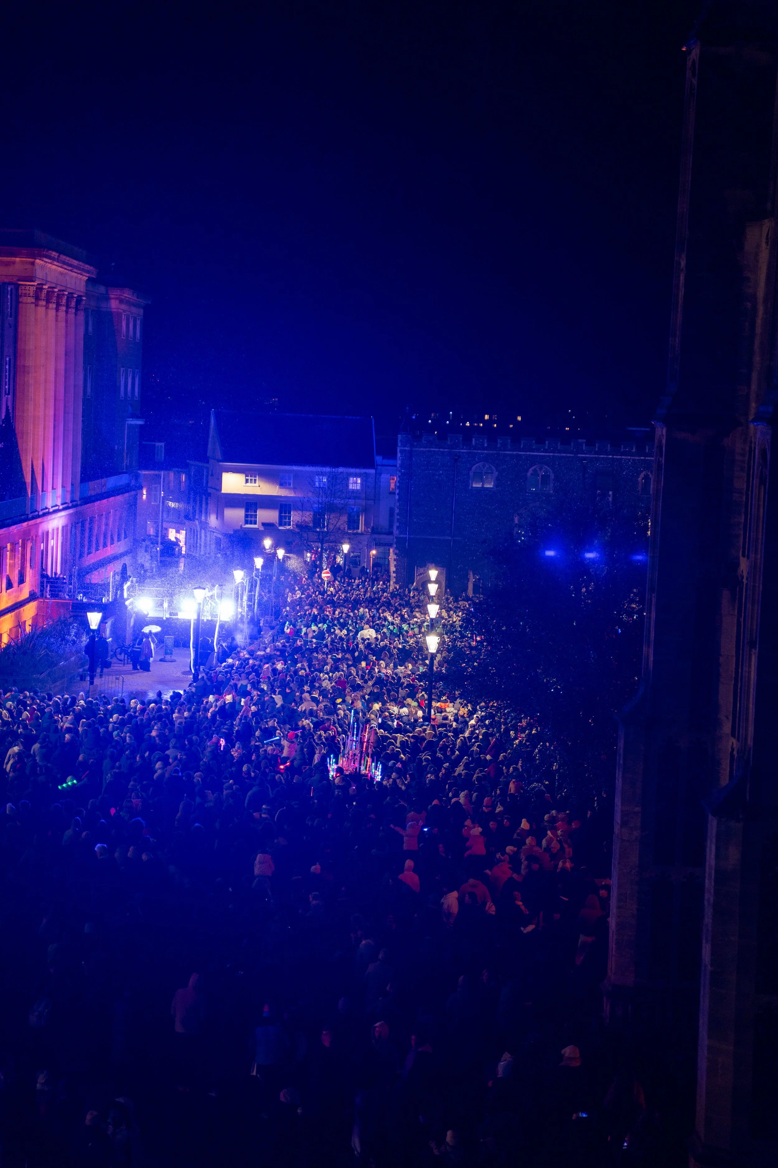 Nighttime scene of a large crowd gathered in a city square illuminated with purple and blue lighting, with historic buildings in the background and street lamps lighting the area.