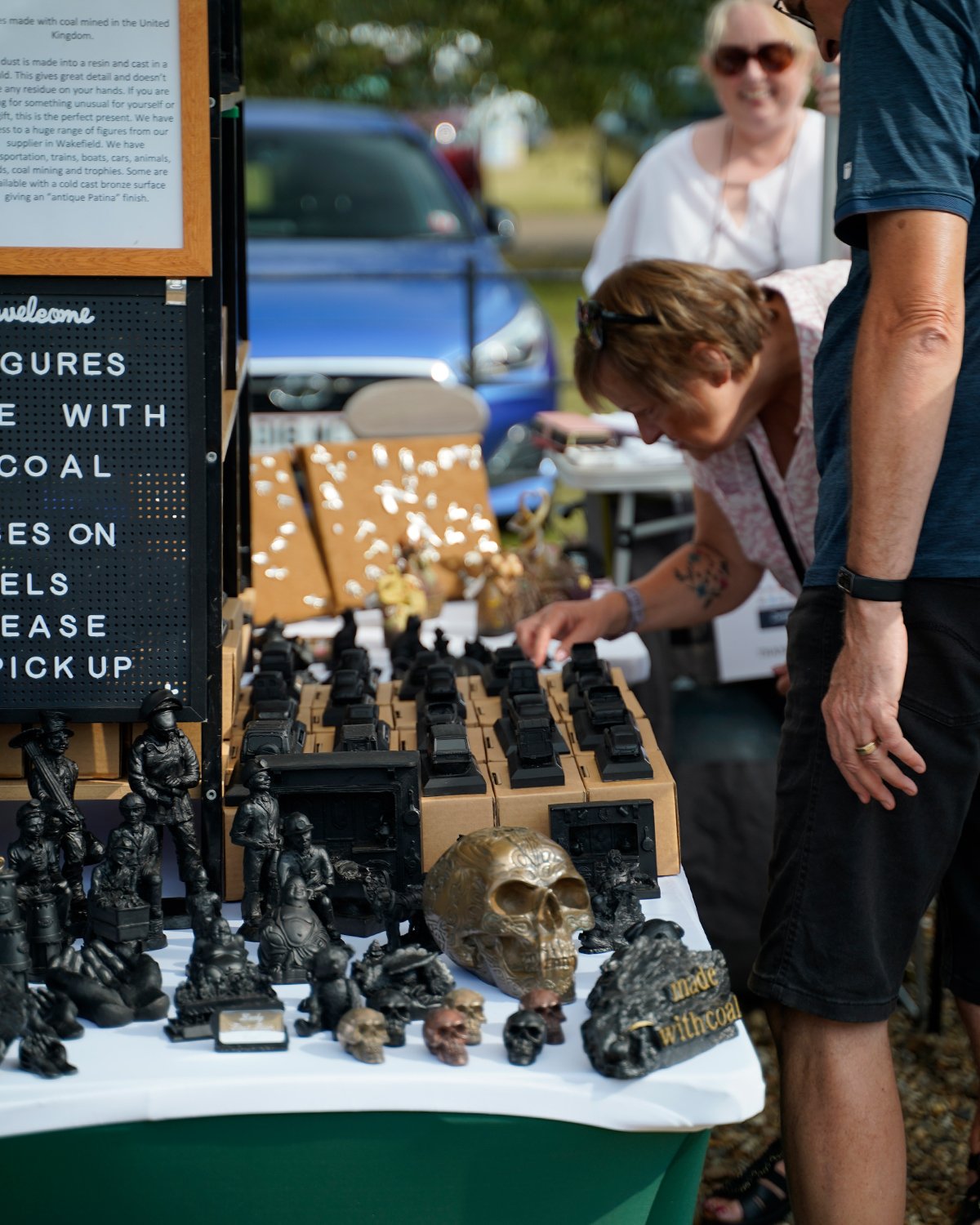 A market stall displaying various black and bronze skull sculptures and figurines, with a person examining the items. A woman is in the background, smiling and looking at the scene, with cars parked behind them and a signboard to the left.