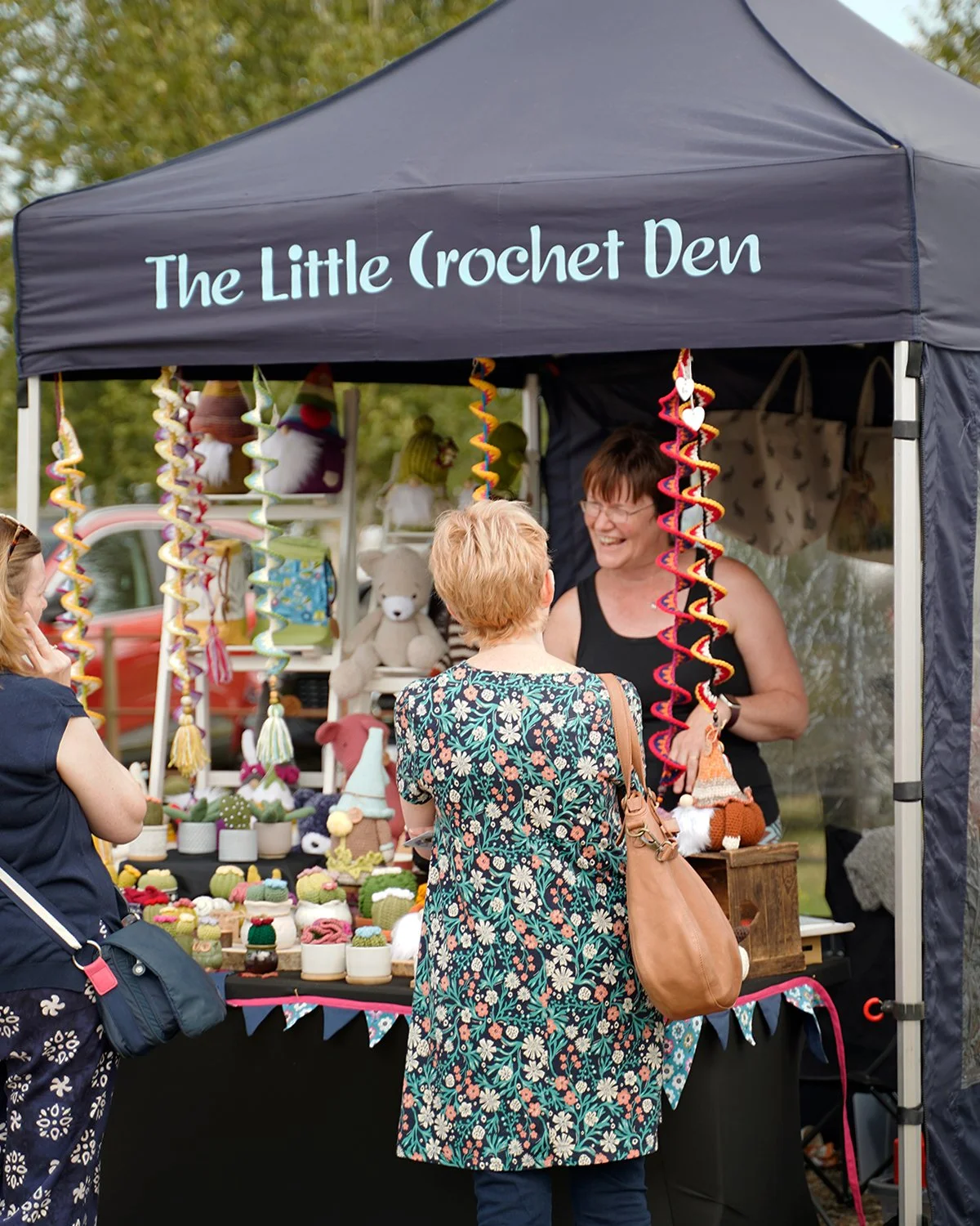 Women are shopping at a craft stall named 'The Little Crochet Den' at an outdoor market, with handmade crochet items and toys on display.