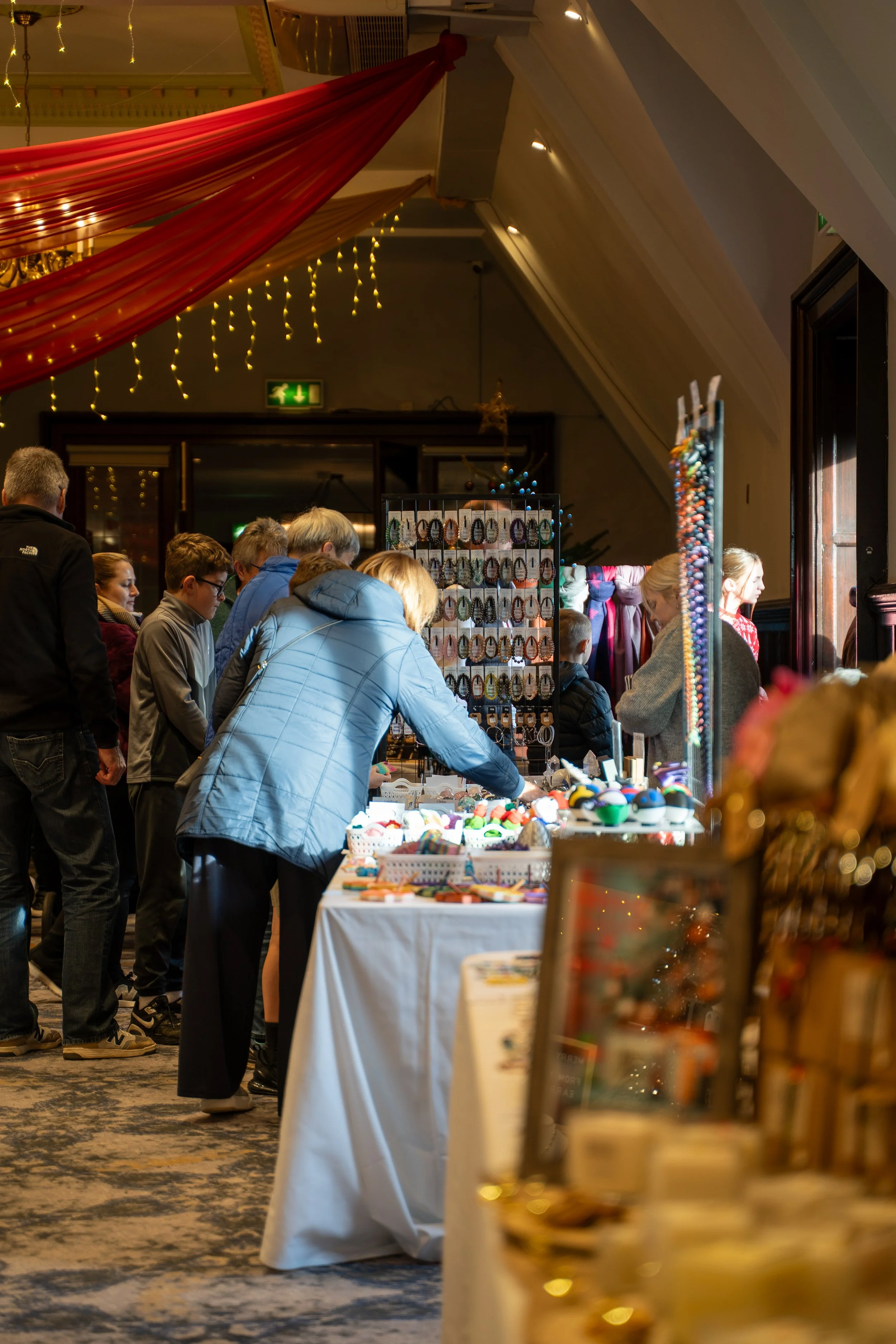 People shopping at an indoor market stall selling jewelry and handmade crafts, with lights and decorations in the background.