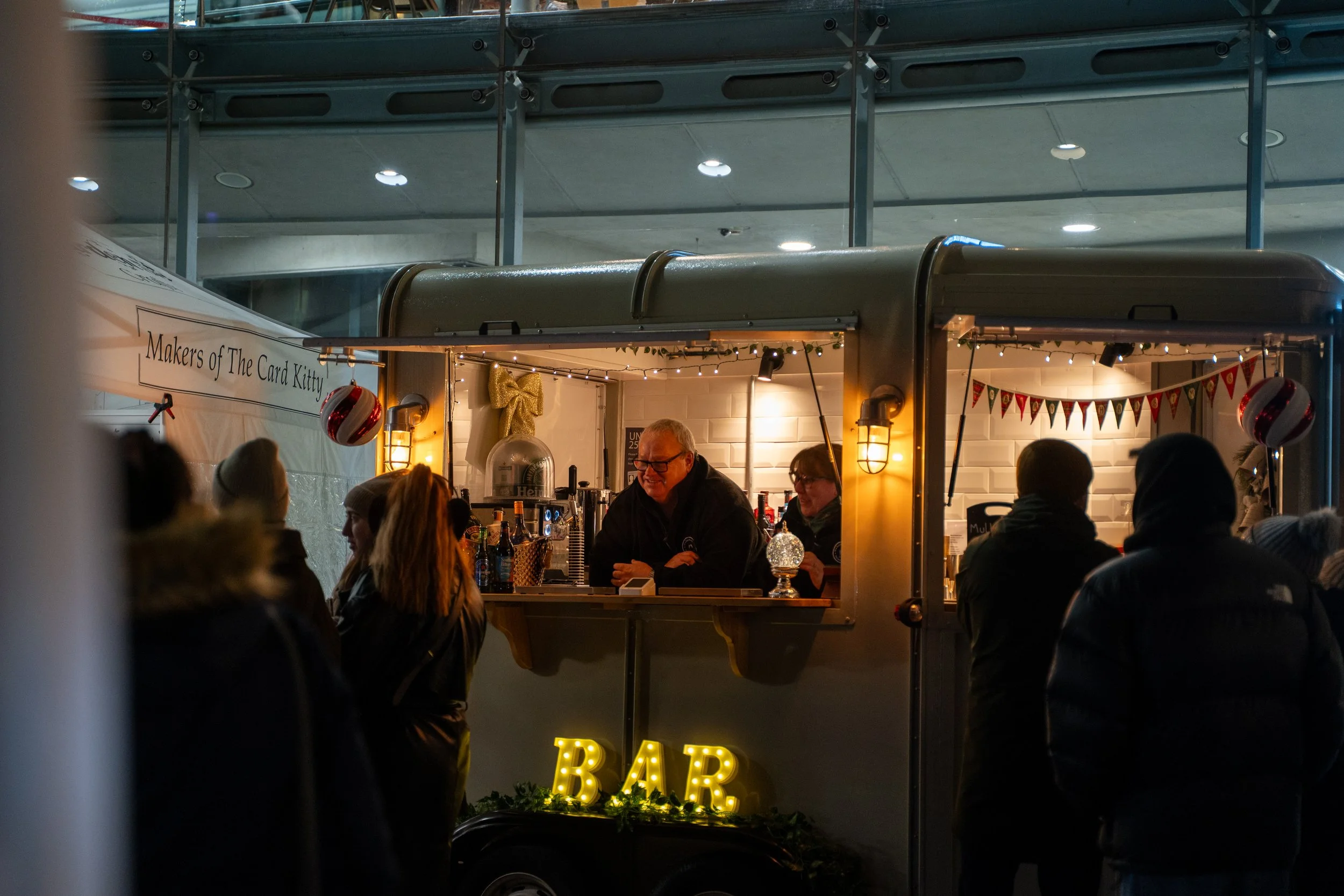 A nighttime scene at an outdoor bar with a food truck setup. The bar is decorated with string lights, Christmas ornaments, and a glowing 'BAR' sign. People are gathered around the bar area, engaging with the bartender, under a roof decorated with a pennant banner.
