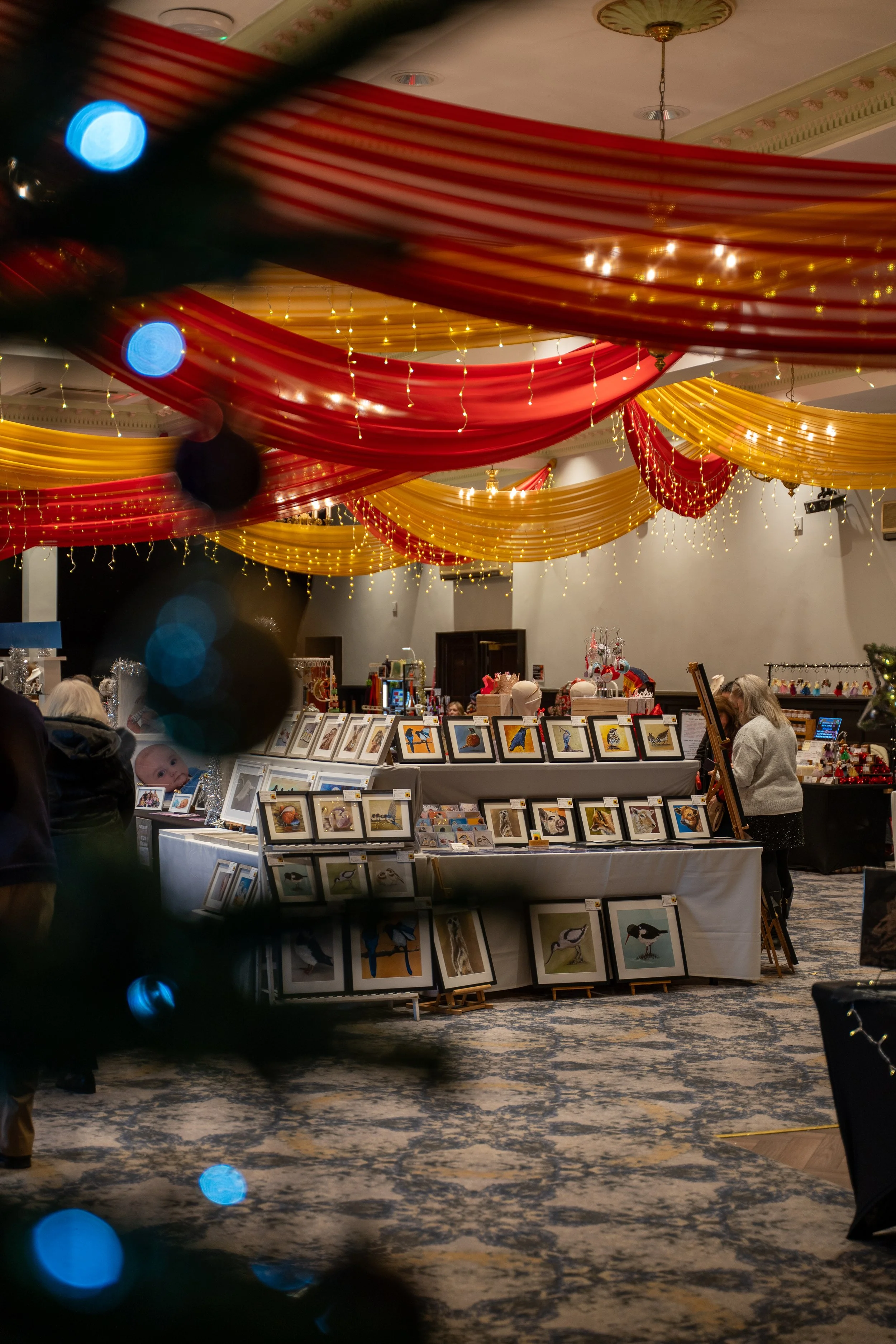 Indoor art fair with paintings of birds on display, decorated with red and yellow draped fabric and string lights, with people browsing the artwork.