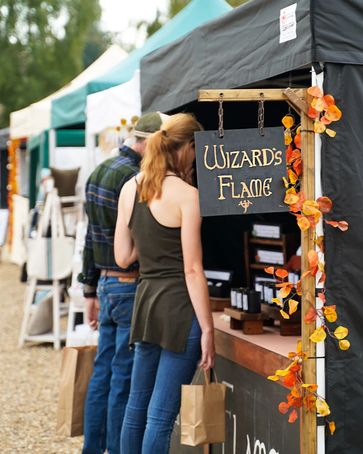 People shopping at a booth labeled "Wizard's Flame" with autumn decorations at an outdoor market.
