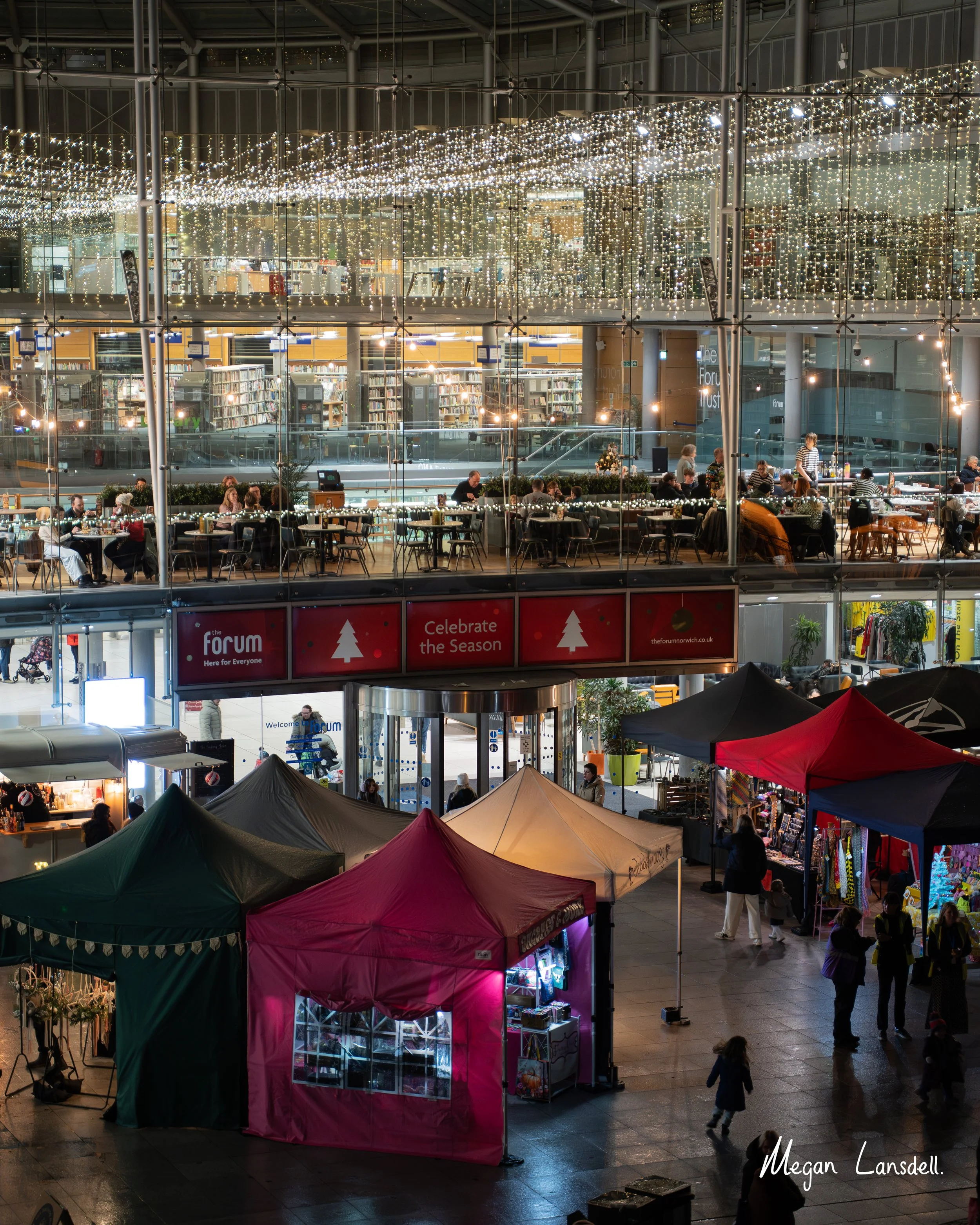 Inside a shopping mall, decorated with string lights hanging from the ceiling. There are multiple small market stalls set up with tents, selling various goods. People are shopping and walking through the area, and a large bookshelf display is visible