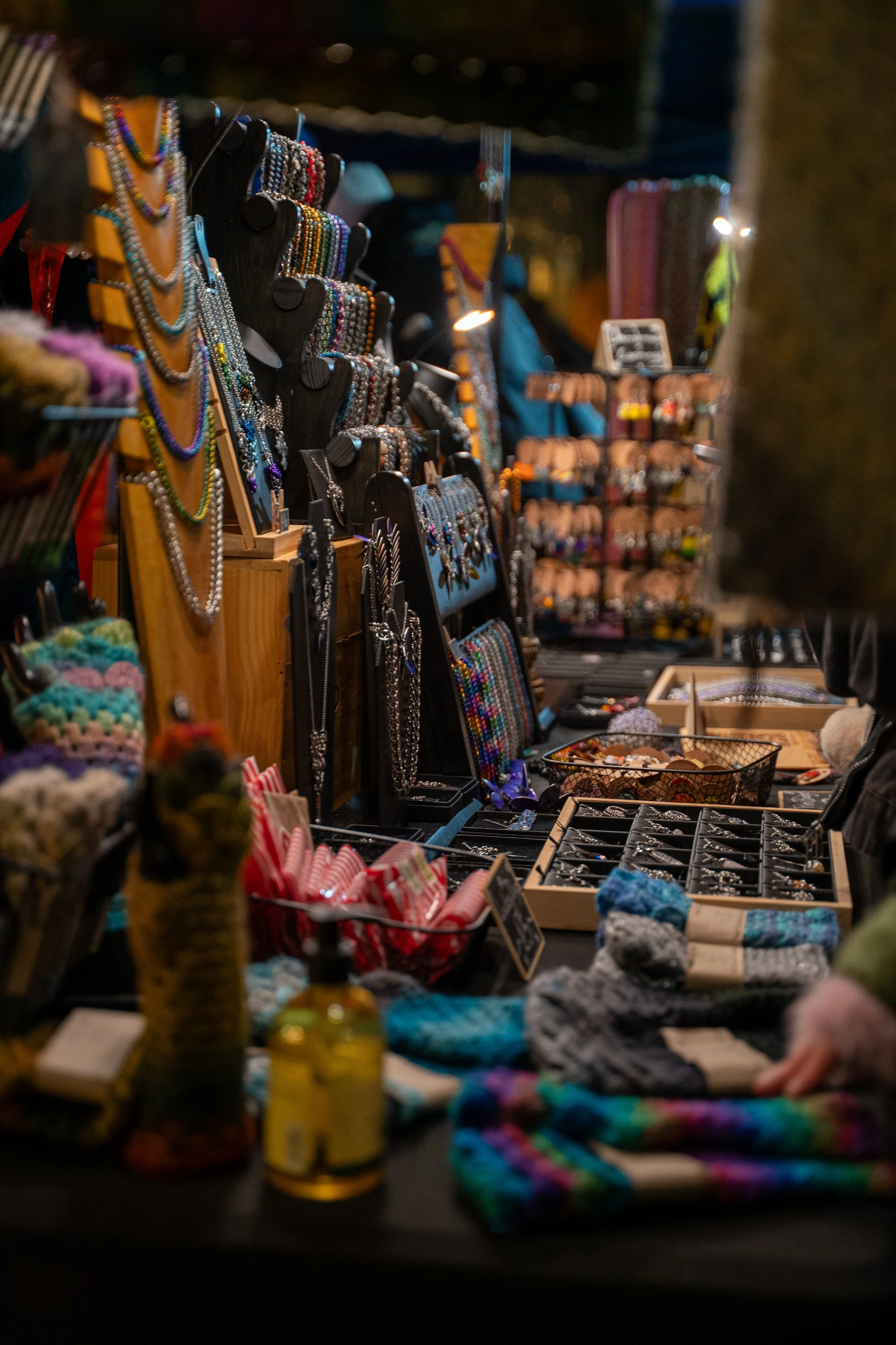 A marketplace stall displaying handmade jewelry, jewelry stands with necklaces and bracelets, and various other accessories including knitted items and bottles.